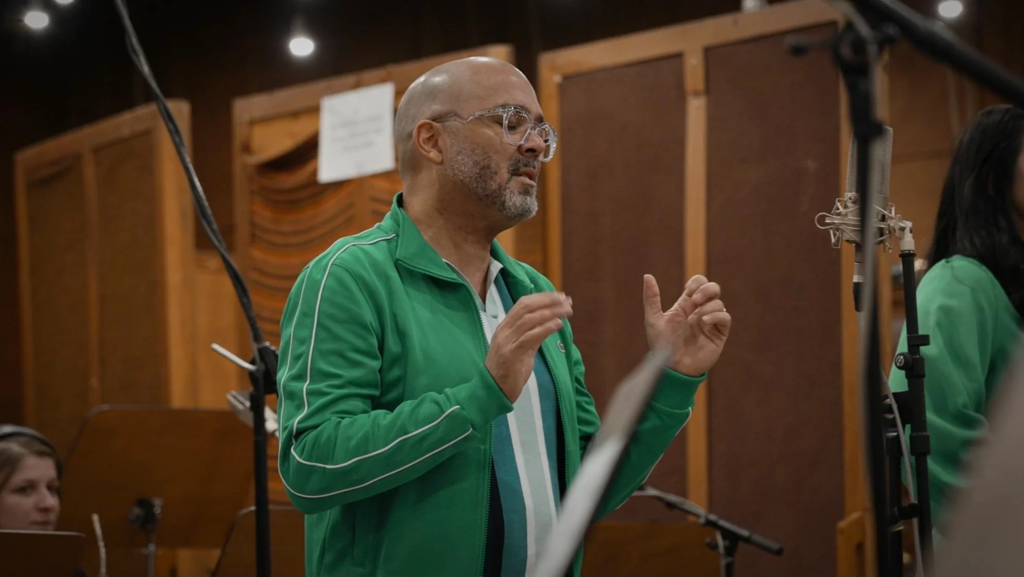 Hombre con gafas y barba, vestido con sudadera verde, dirigiéndose a un micrófono en un estudio de grabación.