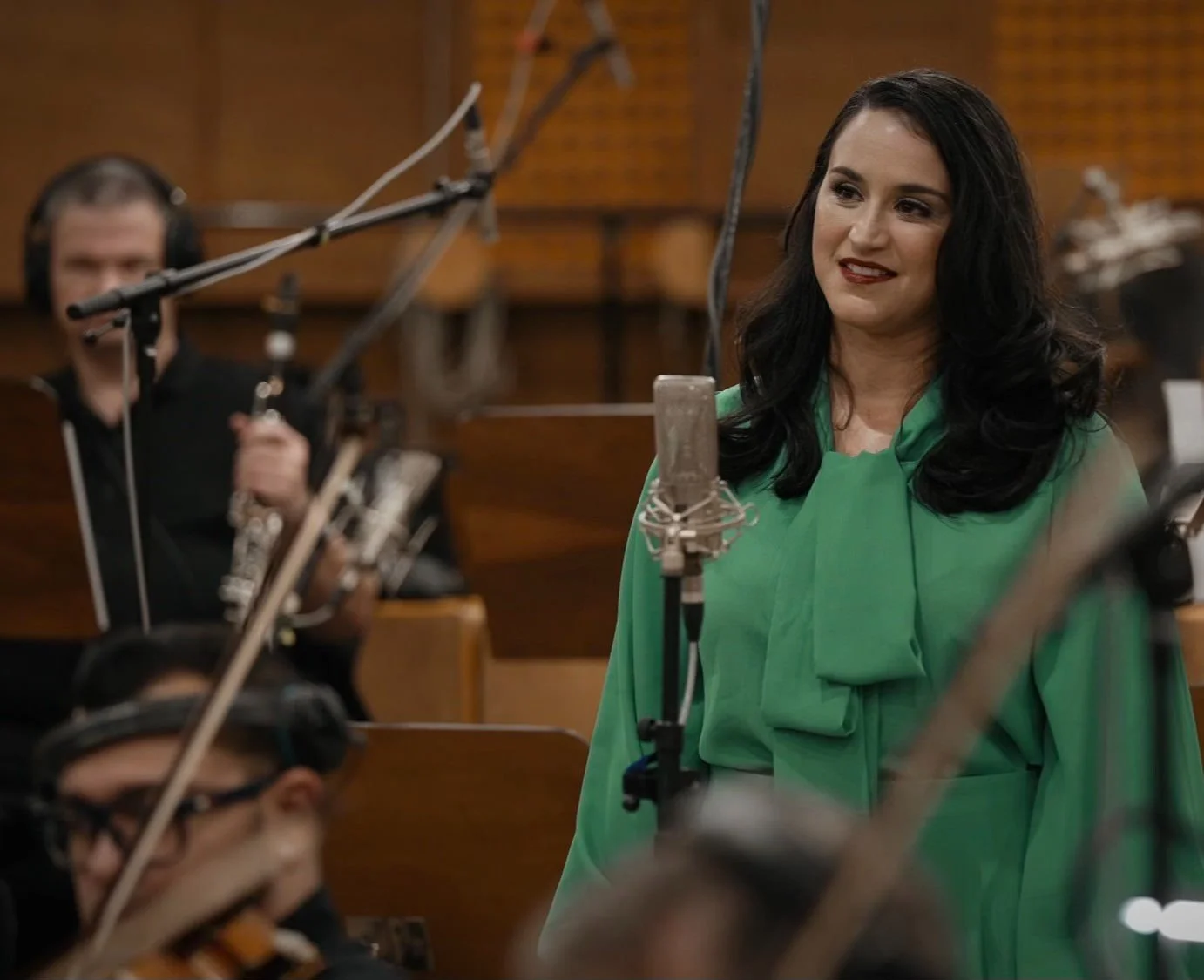 Una mujer con cabello oscuro y largo, vestida con un traje verde, en un estudio de grabación rodeada de micrófonos y músicos que tocan instrumentos de cuerda.