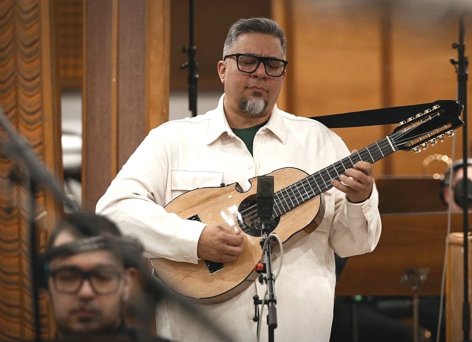 Hombre tocando guitarra en un estudio de grabación, con cables y otros músicos alrededor.