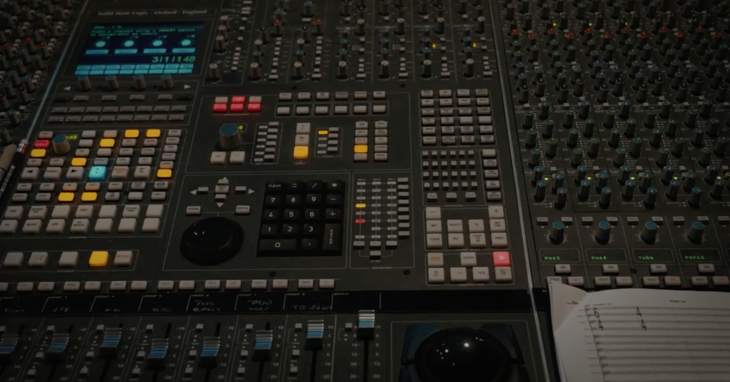 Vista de una consola de mezclas de audio con múltiples botones y perillas, y una hoja de papel con notas musicales a la derecha.