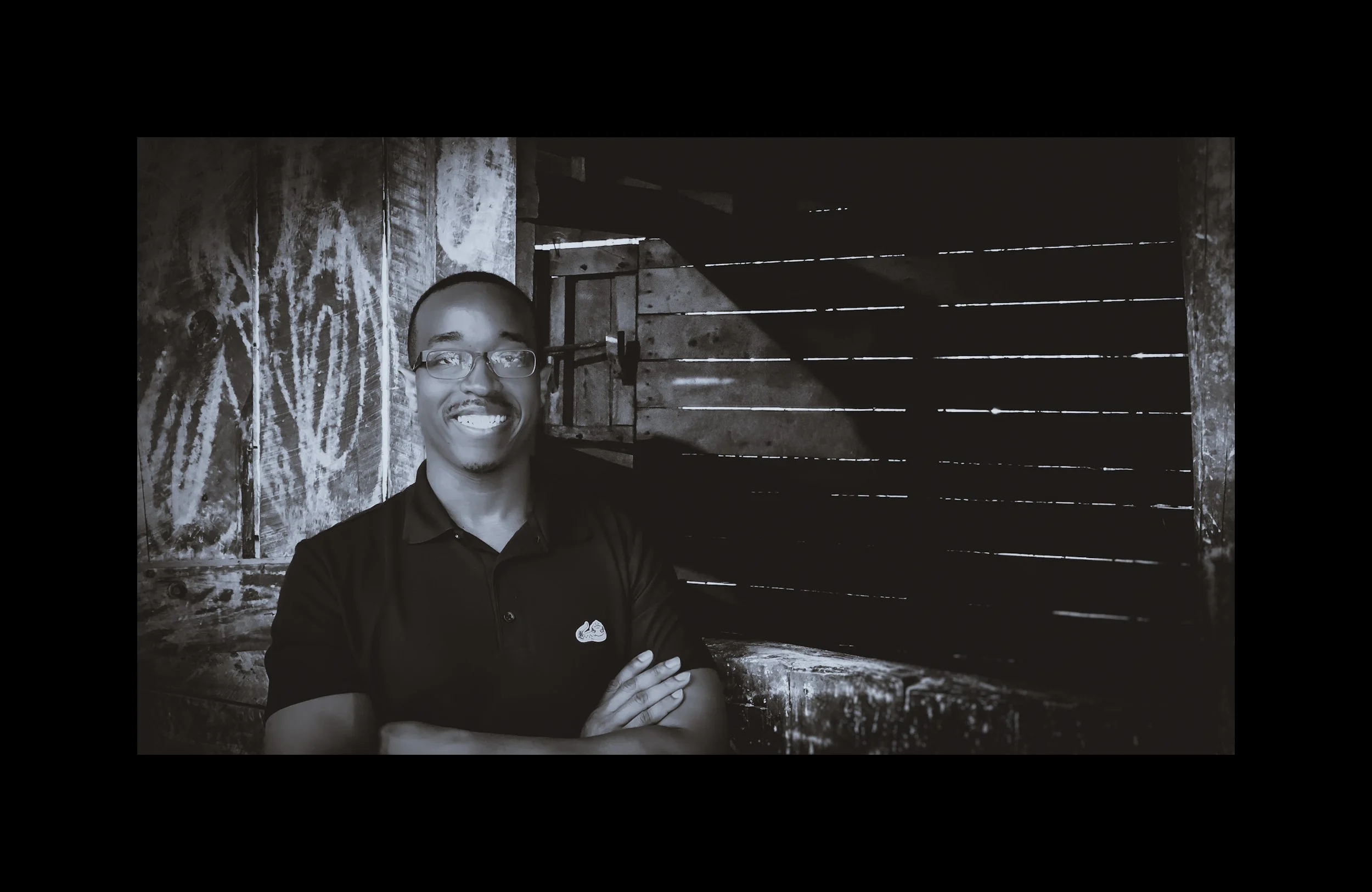 A man with glasses, smiling, wearing a black collared shirt with a small logo, standing with arms crossed in front of a rustic wooden wall and door.