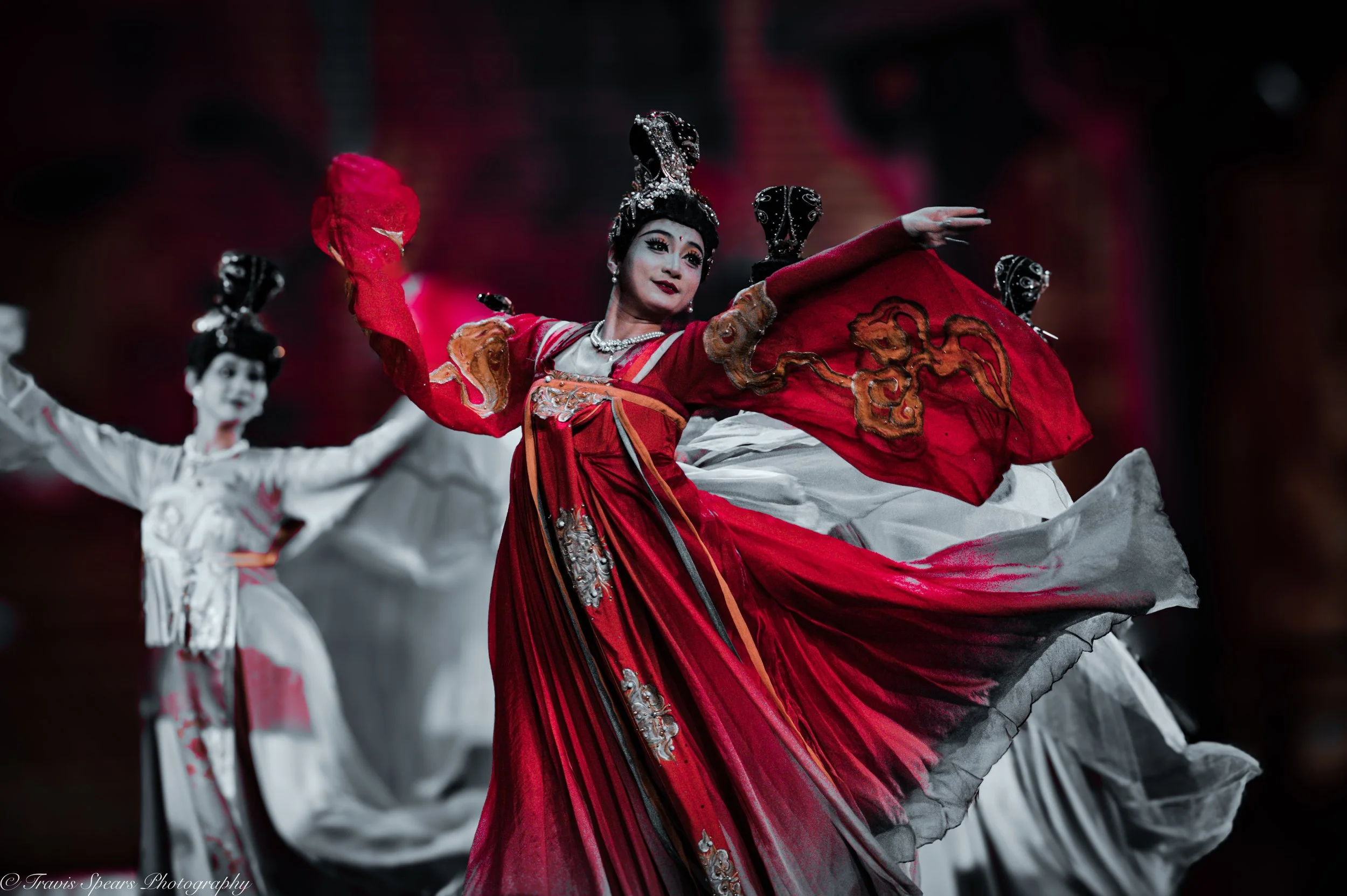 Dancer dressed in a red and white traditional costume performing a dance with flowing fabric, accompanied by other dancers in white costumes.
