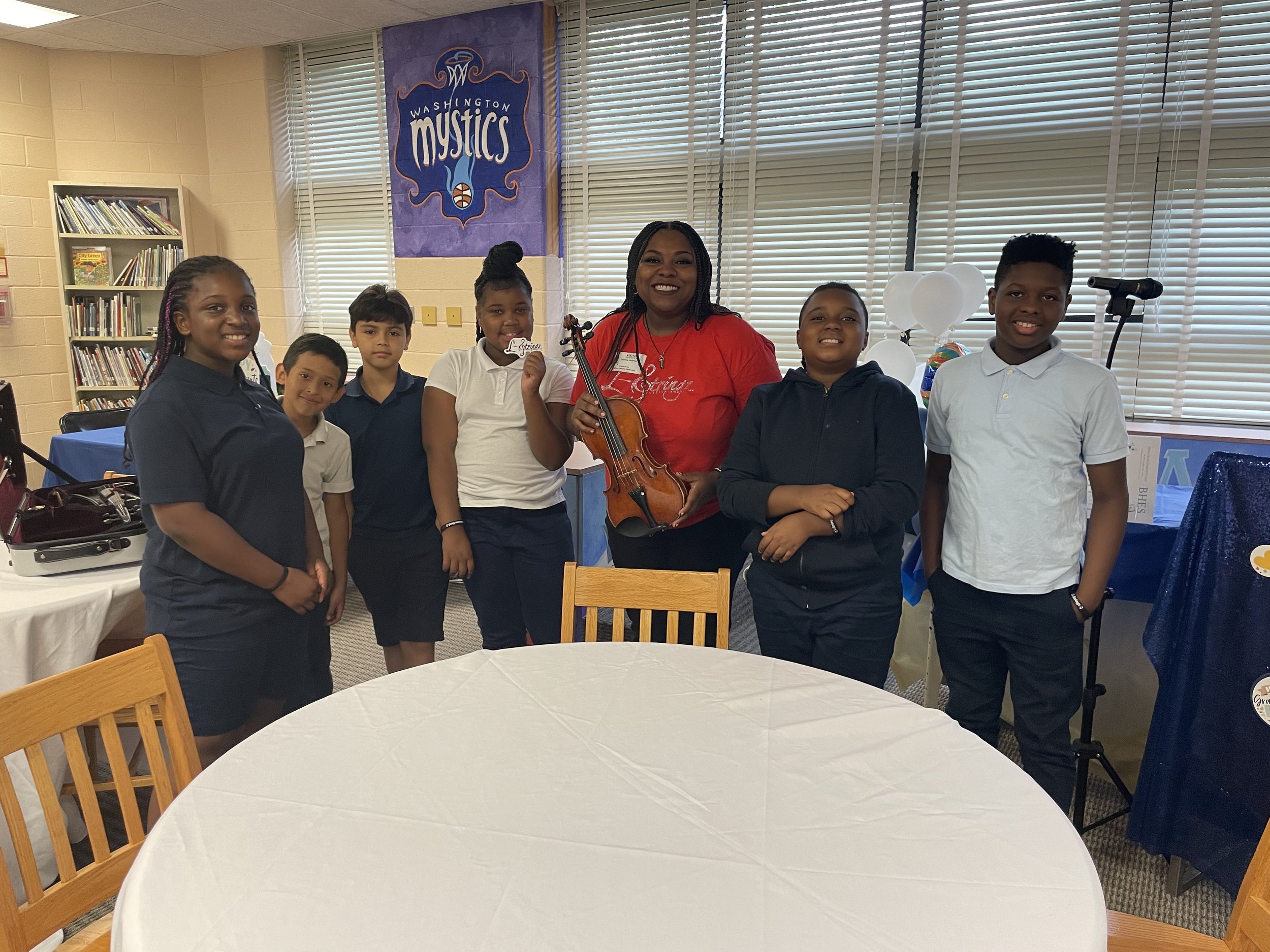 Group of six children and a woman holding a violin in a classroom. The children are smiling, and the classroom has bookshelves, balloons, and a Washington Mystics banner in the background.