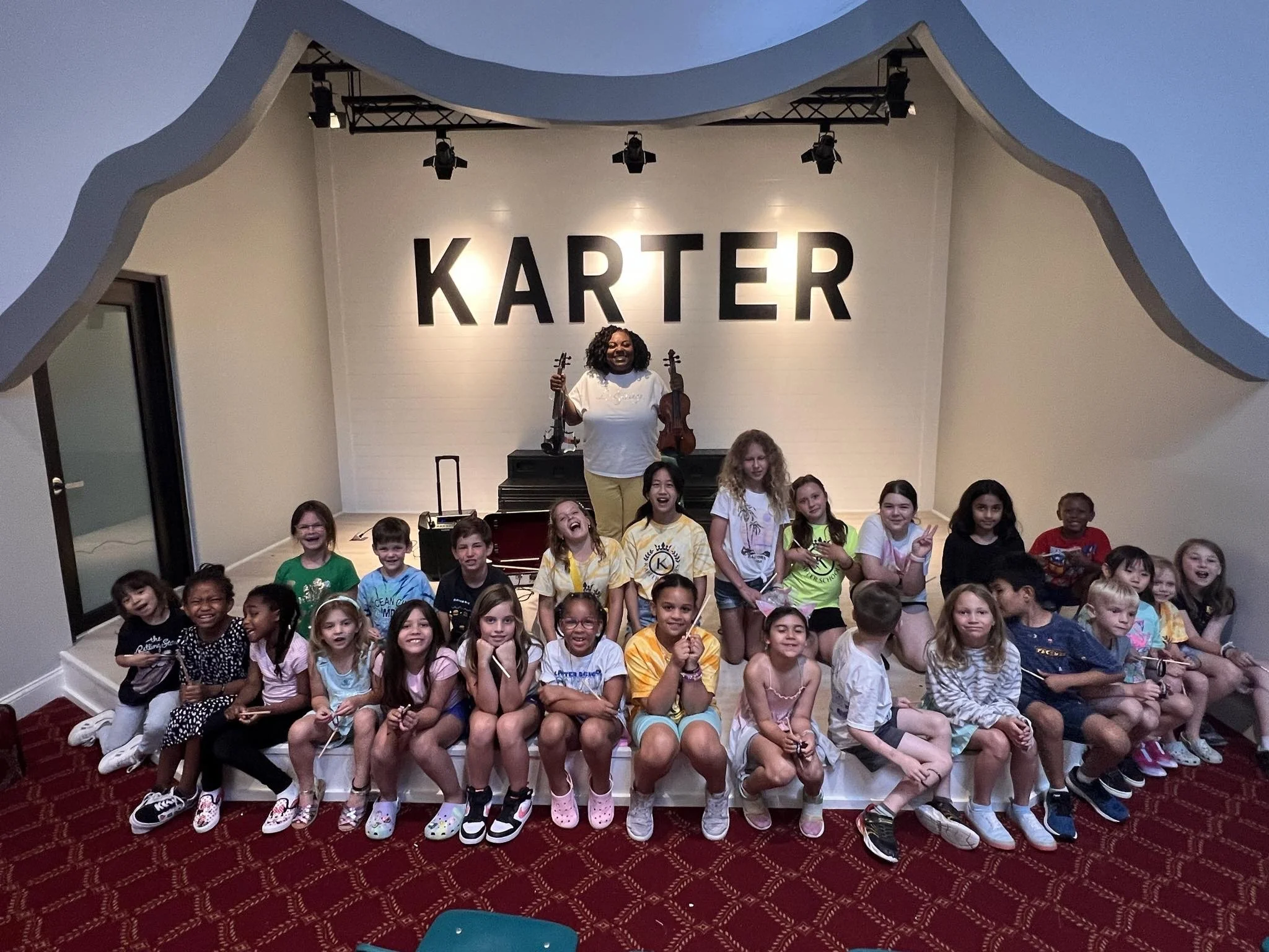 Group of children sitting and standing on a stage with a woman holding a violin, in front of a large illuminated sign that says "KARTER" at an indoor event.