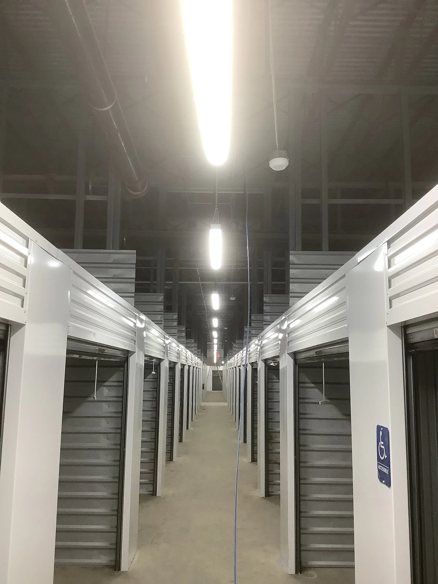Long hallway with rows of small office or storage units with open doors, bright overhead fluorescent lighting, and a sign indicating accessible entry on the right side.