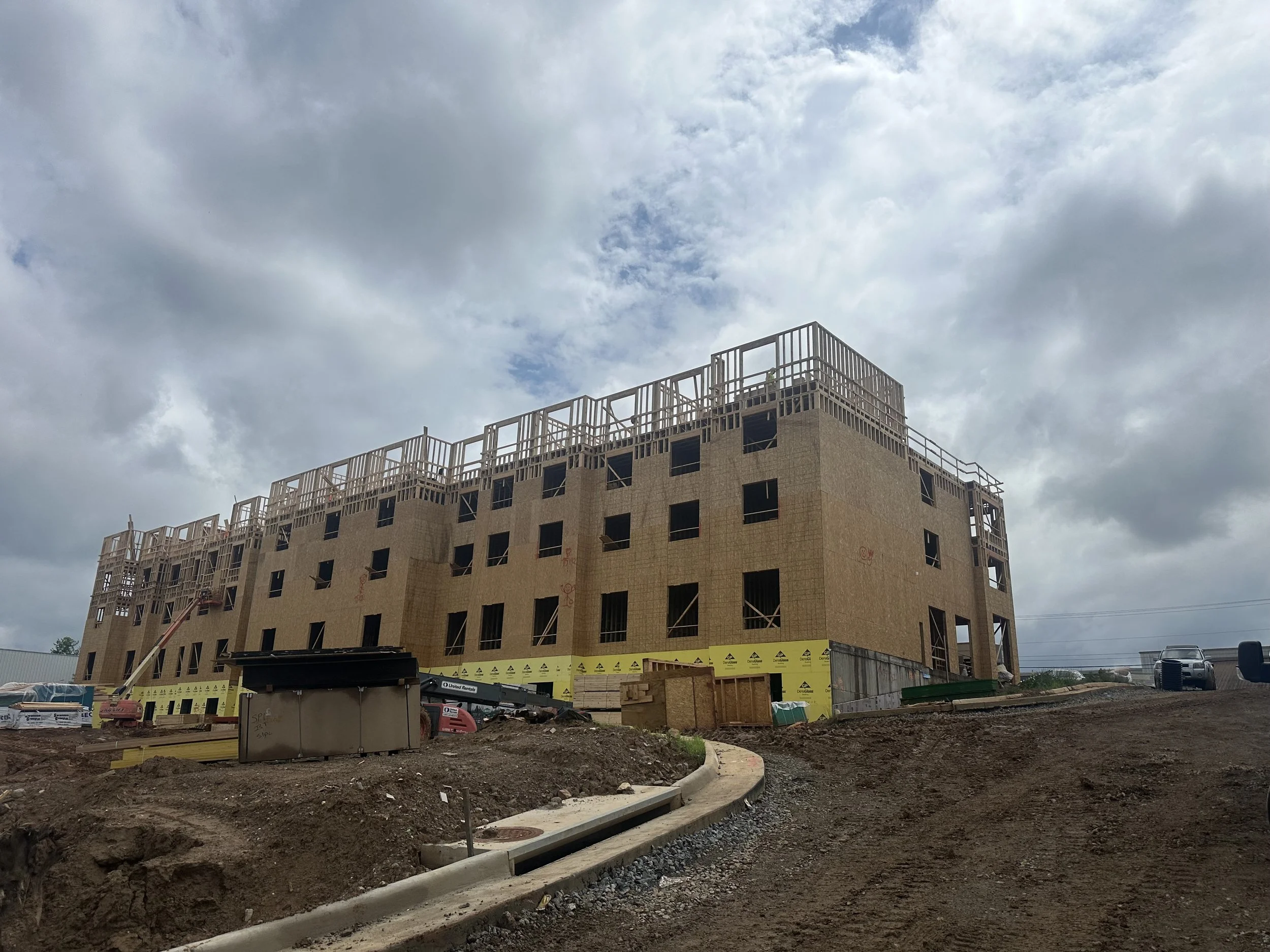 Construction worker standing next to a dug trench with several metal pipes laid inside, a measuring tape indicating approximately 21 inches tall, and construction tools nearby.