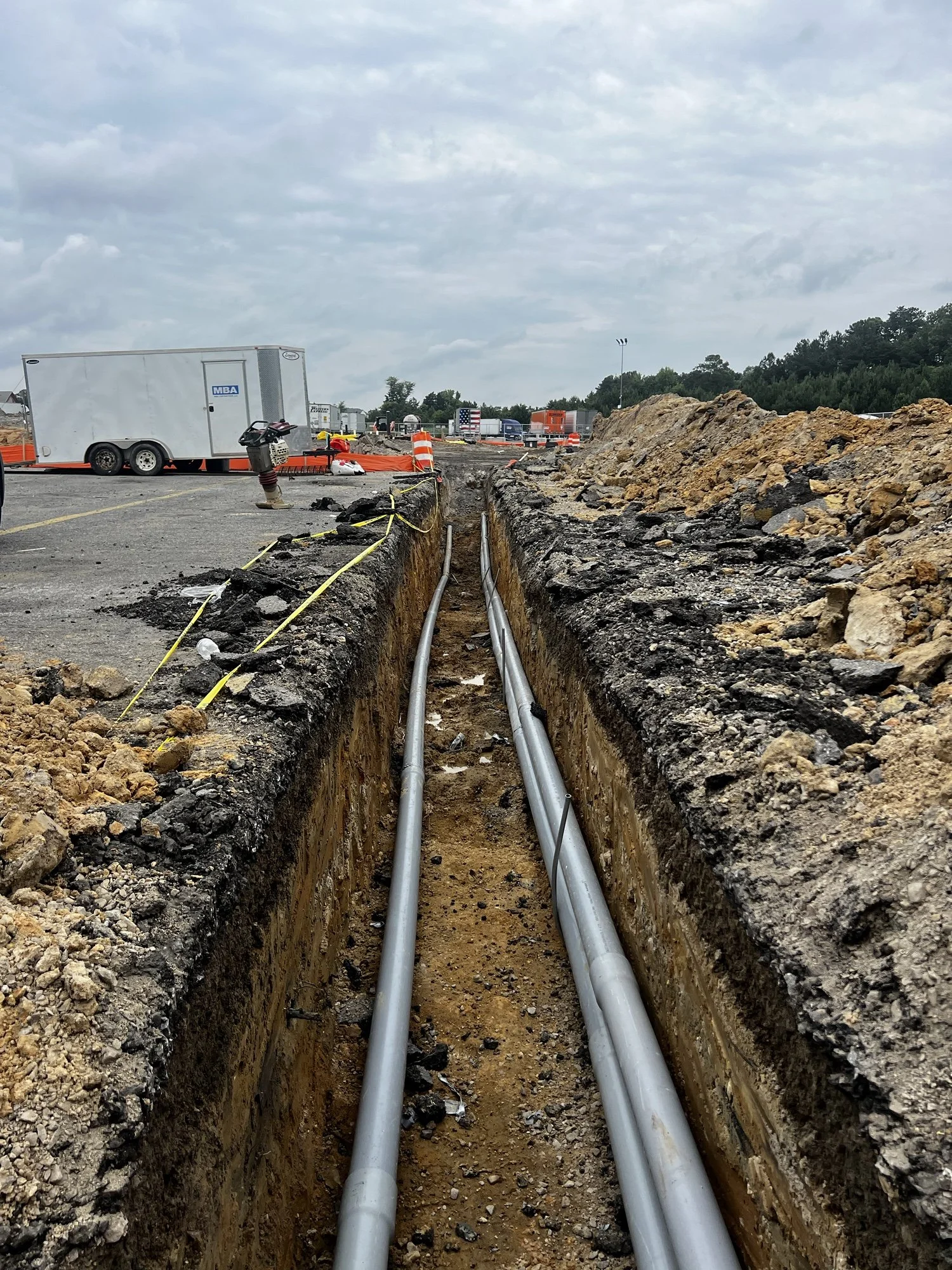 Construction site with excavation trench and utility pipes, trailers and construction equipment in background.
