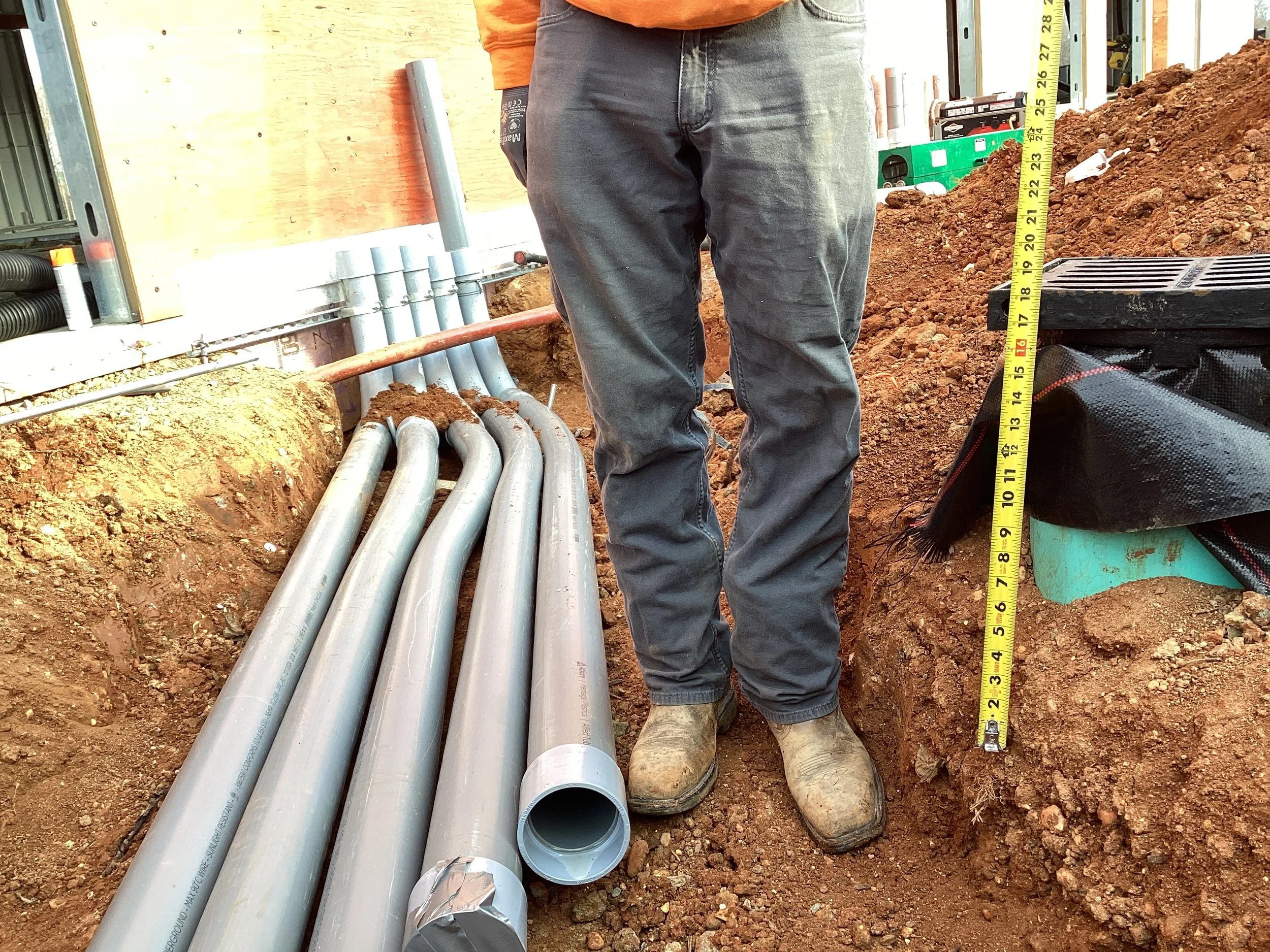 Construction worker standing next to a trench with multiple gray pipes on the ground and a measuring tape indicating height.