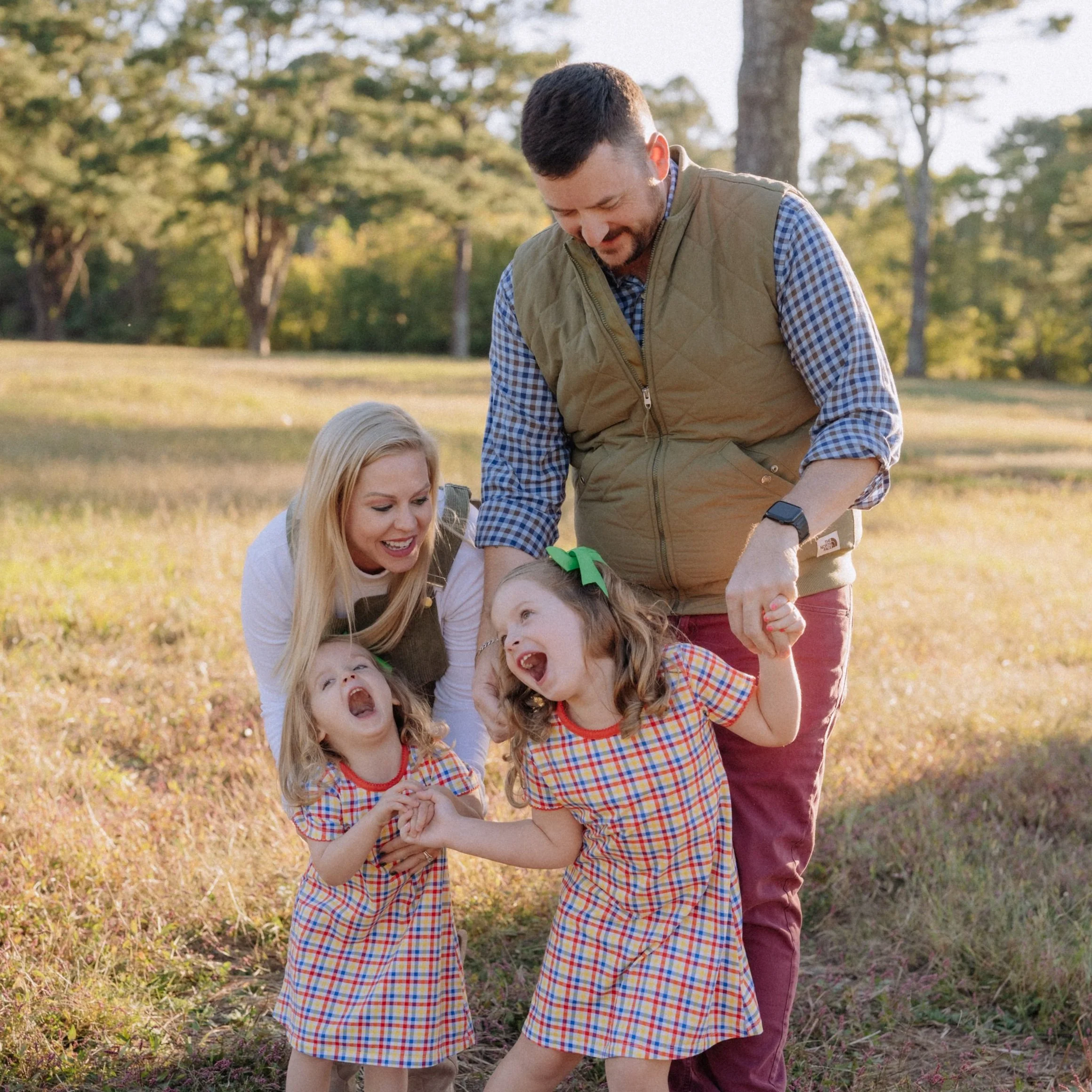 A happy family of four, including two young girls and two adults, enjoying time outdoors in a park with trees. The children are wearing matching plaid dresses, and the adults are smiling and holding their hands.