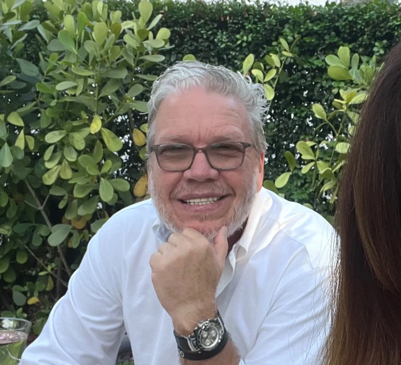 A man with gray hair, glasses, and a beard, wearing a white shirt and a yellow lanyard, smiling in front of a lush tropical garden with large palm leaves and clear blue sky.