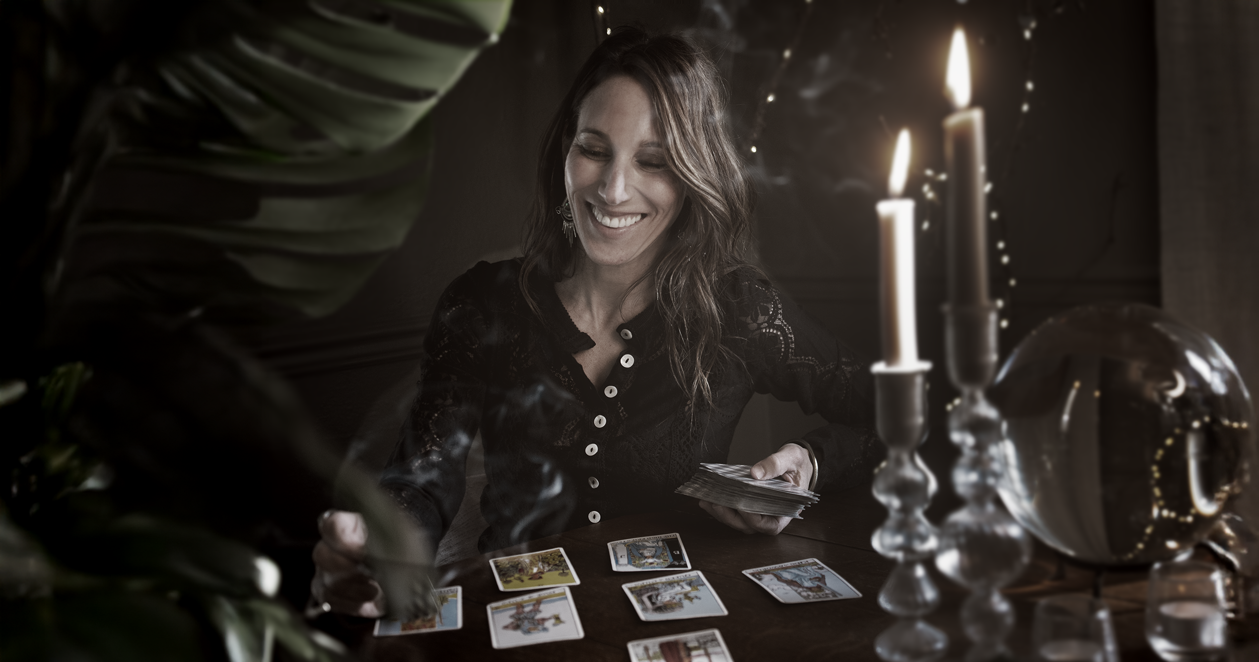 A woman with long wavy hair smiles while sitting at a table covered with tarot cards, holding some cards in her hand. The scene is dimly lit by candles and decorative glass objects, creating a mysterious atmosphere.