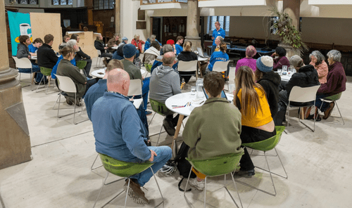 Picture of a people's assembly, showing people sat round tables in a hall