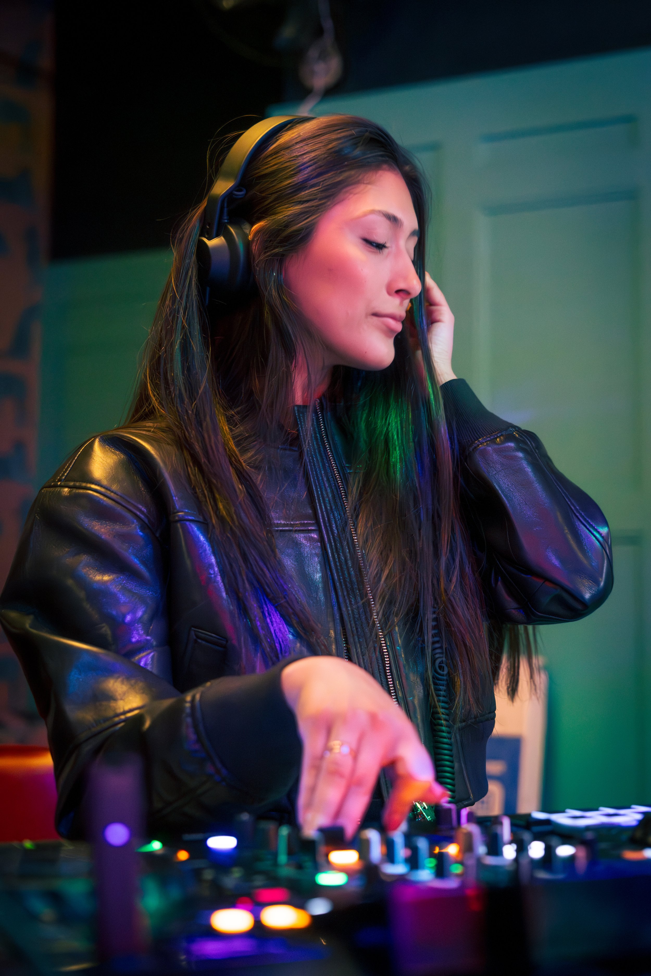Young woman DJ wearing headphones, adjusting mixer controls in a colorful, dimly lit room.