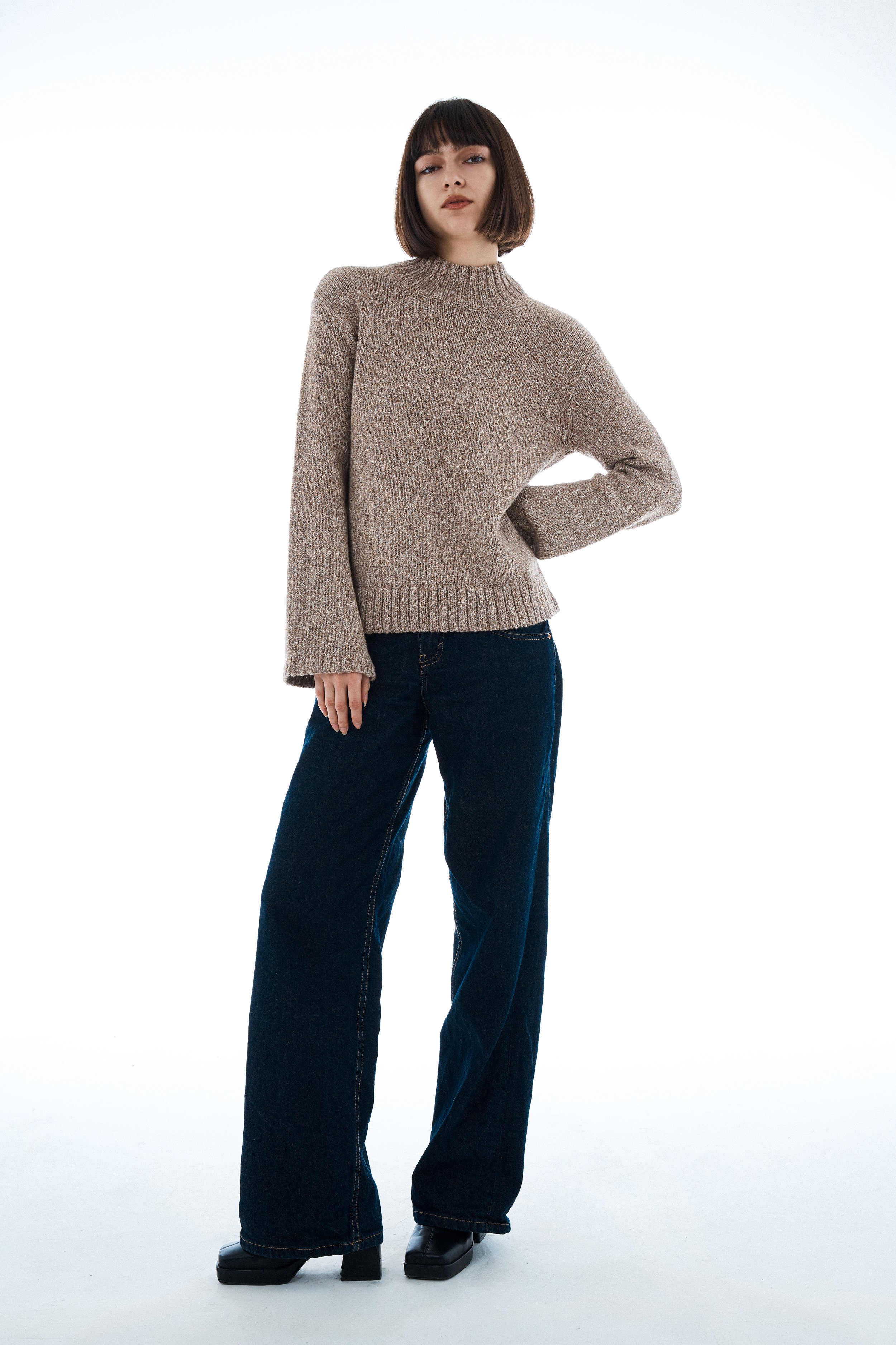 Woman with short dark hair wearing a beige knit sweater and wide-leg dark jeans, standing against a plain white background.