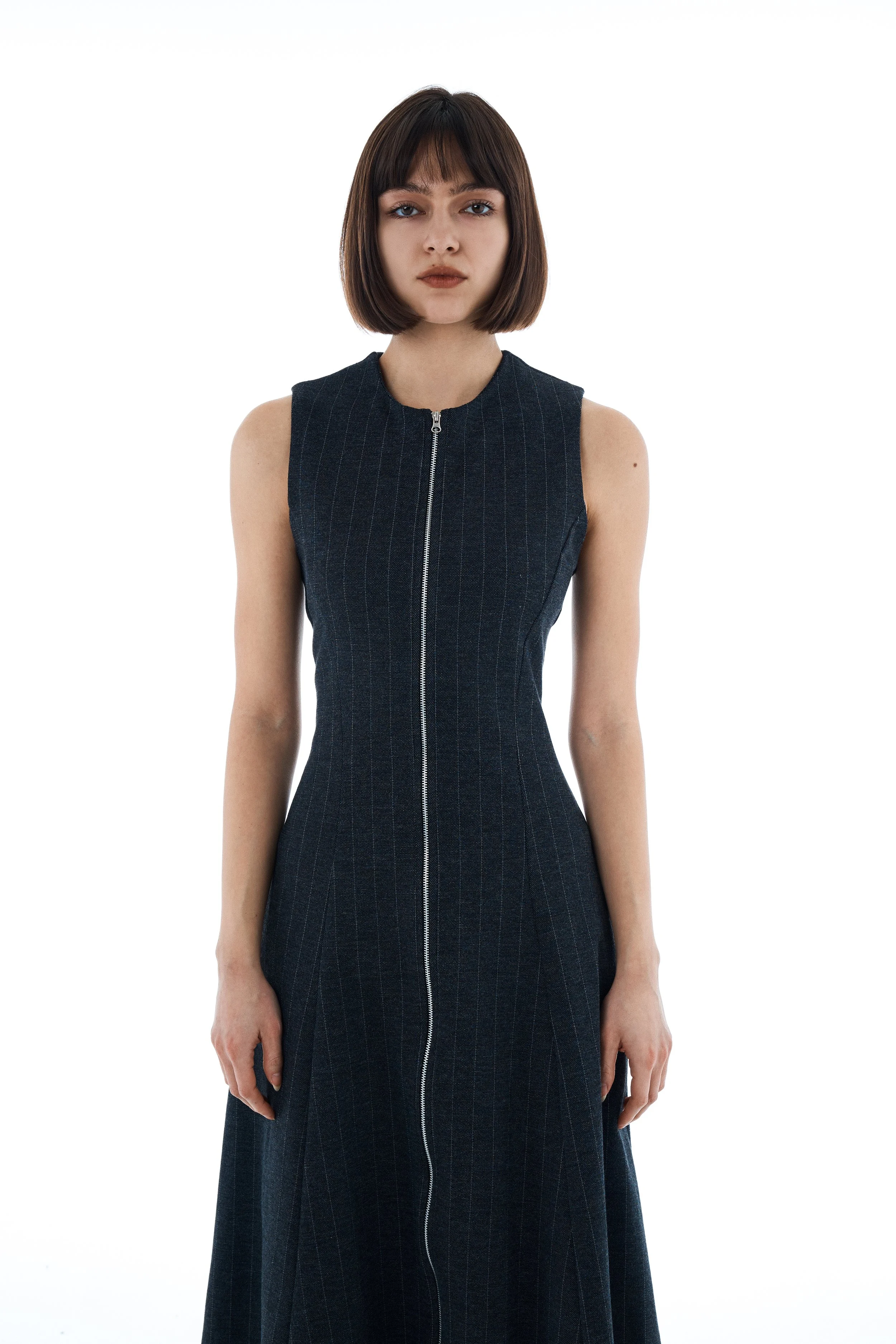 Young woman with shoulder-length brown hair and bangs wearing a sleeveless dark pinstripe zip-up dress, standing against a white background.
