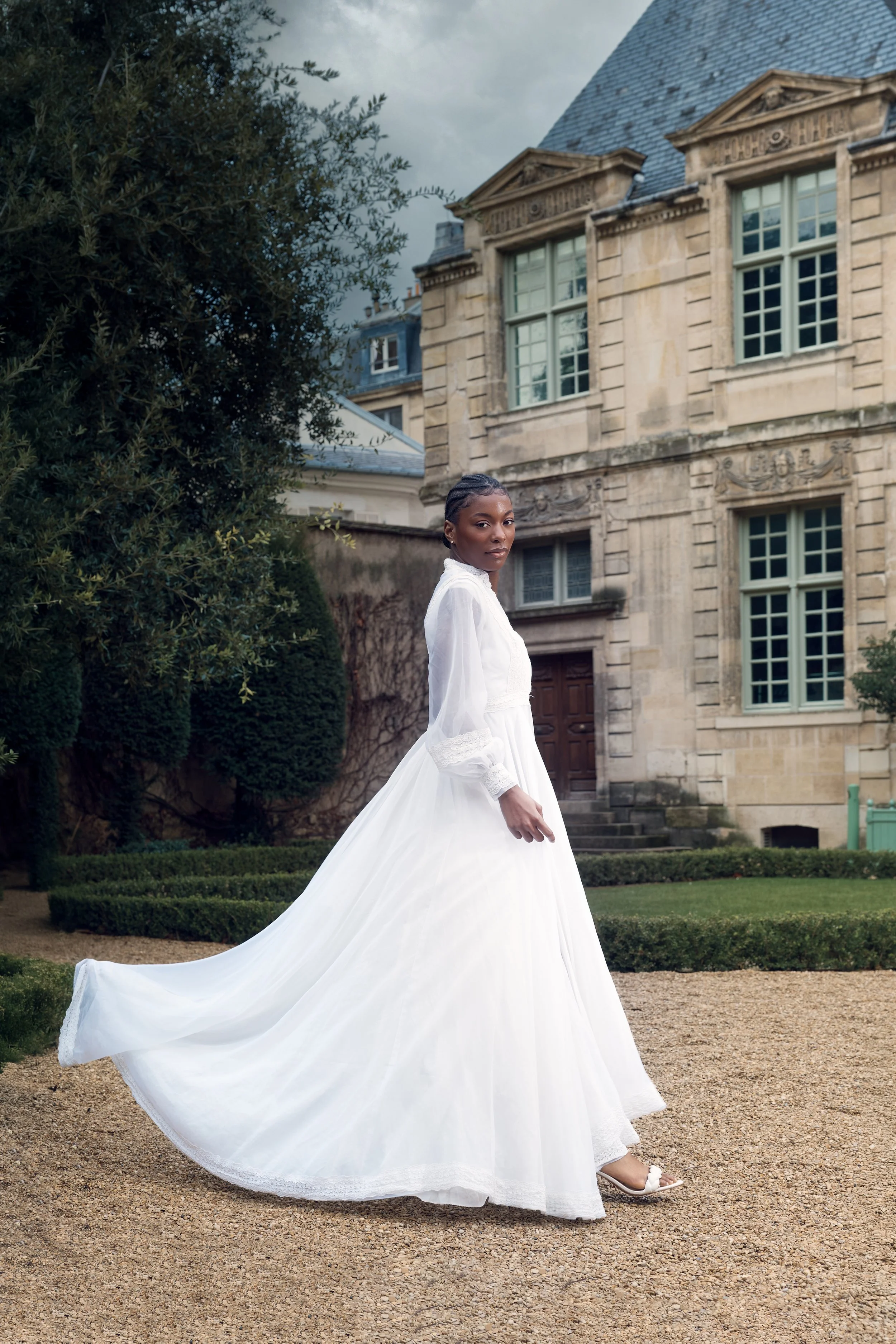A woman in a white wedding dress standing outside in front of an old stone mansion with large windows and a slate roof, surrounded by manicured bushes and trees.