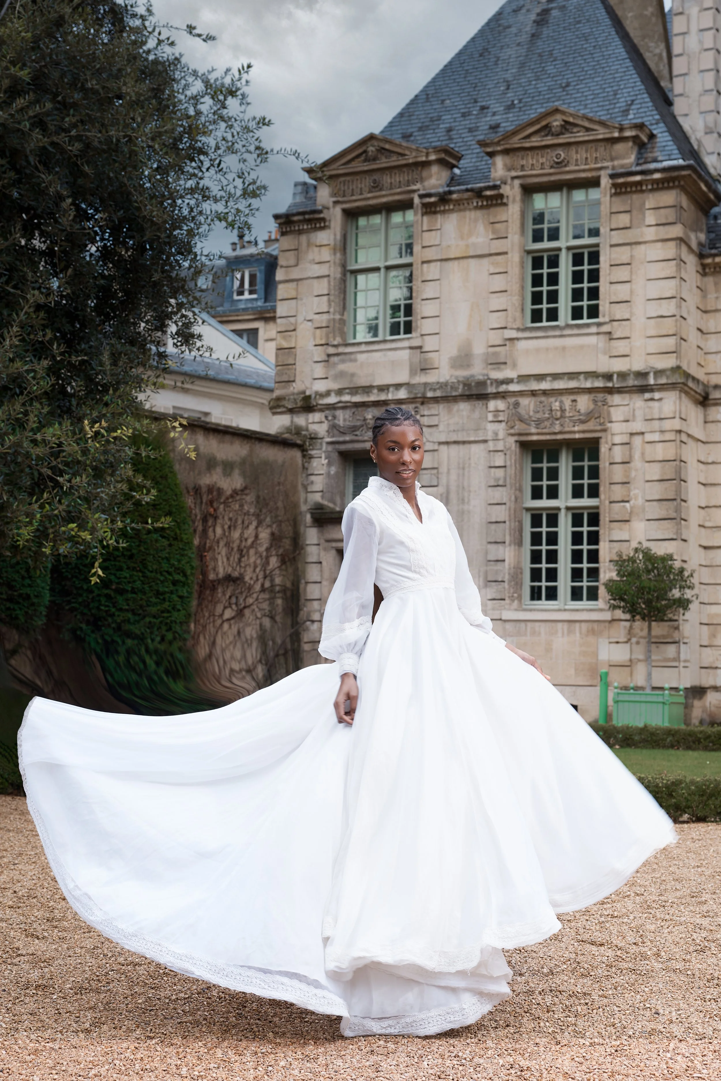 A woman in a white wedding dress standing outdoors in front of a historic stone building with large windows and Gothic architectural details.