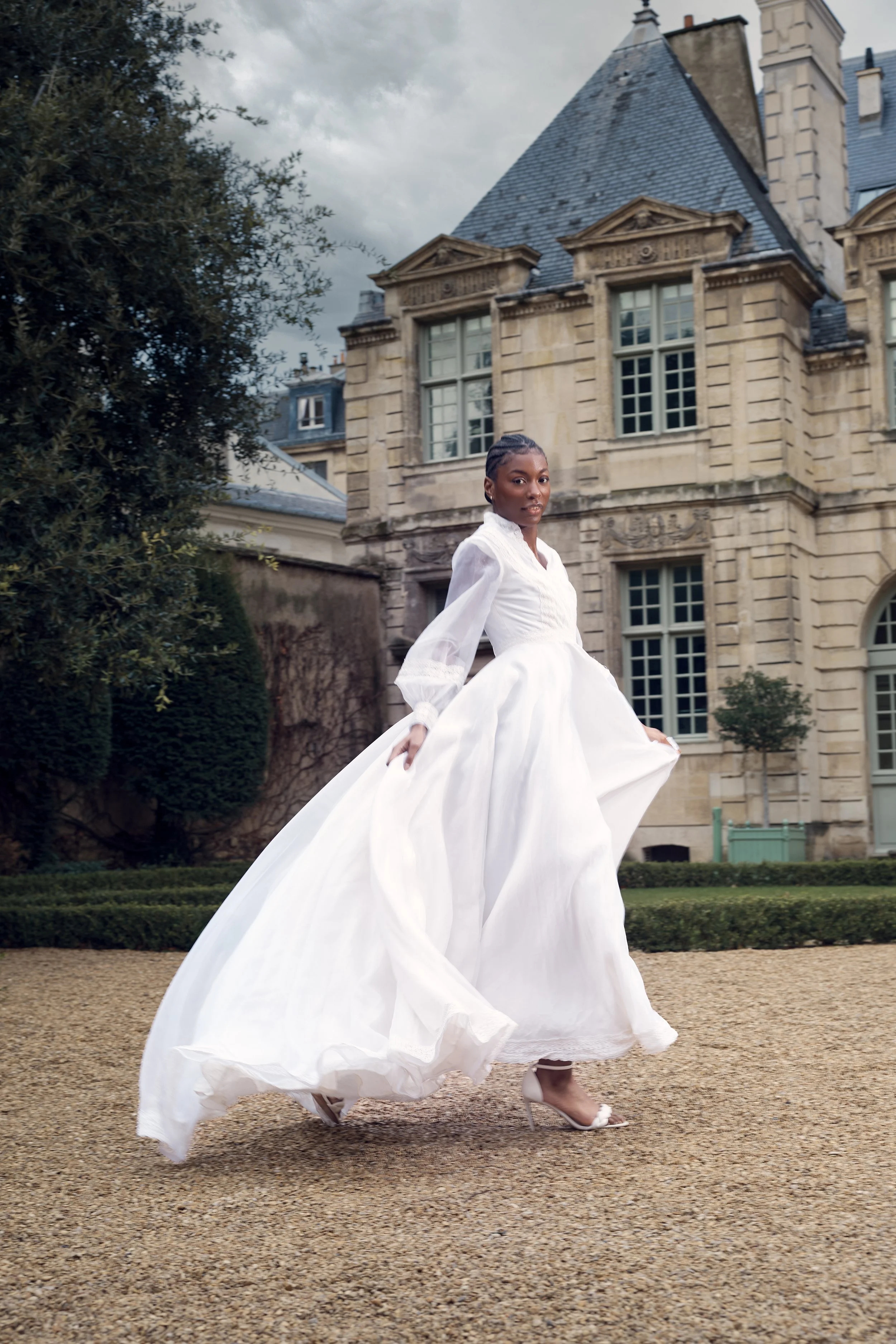 Woman in a white dress and high heels standing outdoors in front of a historic stone mansion.