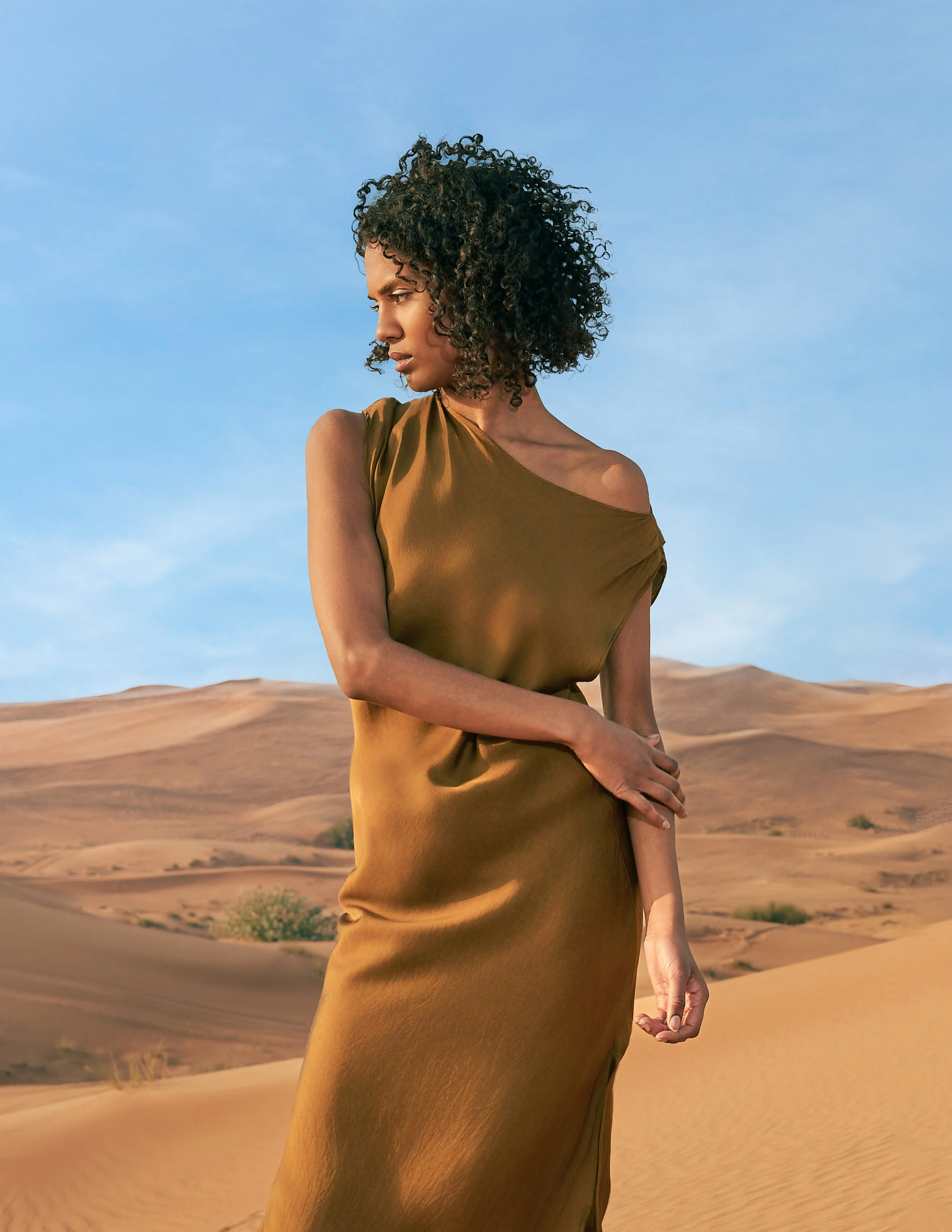 A woman with curly black hair stands in a desert landscape with sand dunes and a clear blue sky, wearing a brown dress.