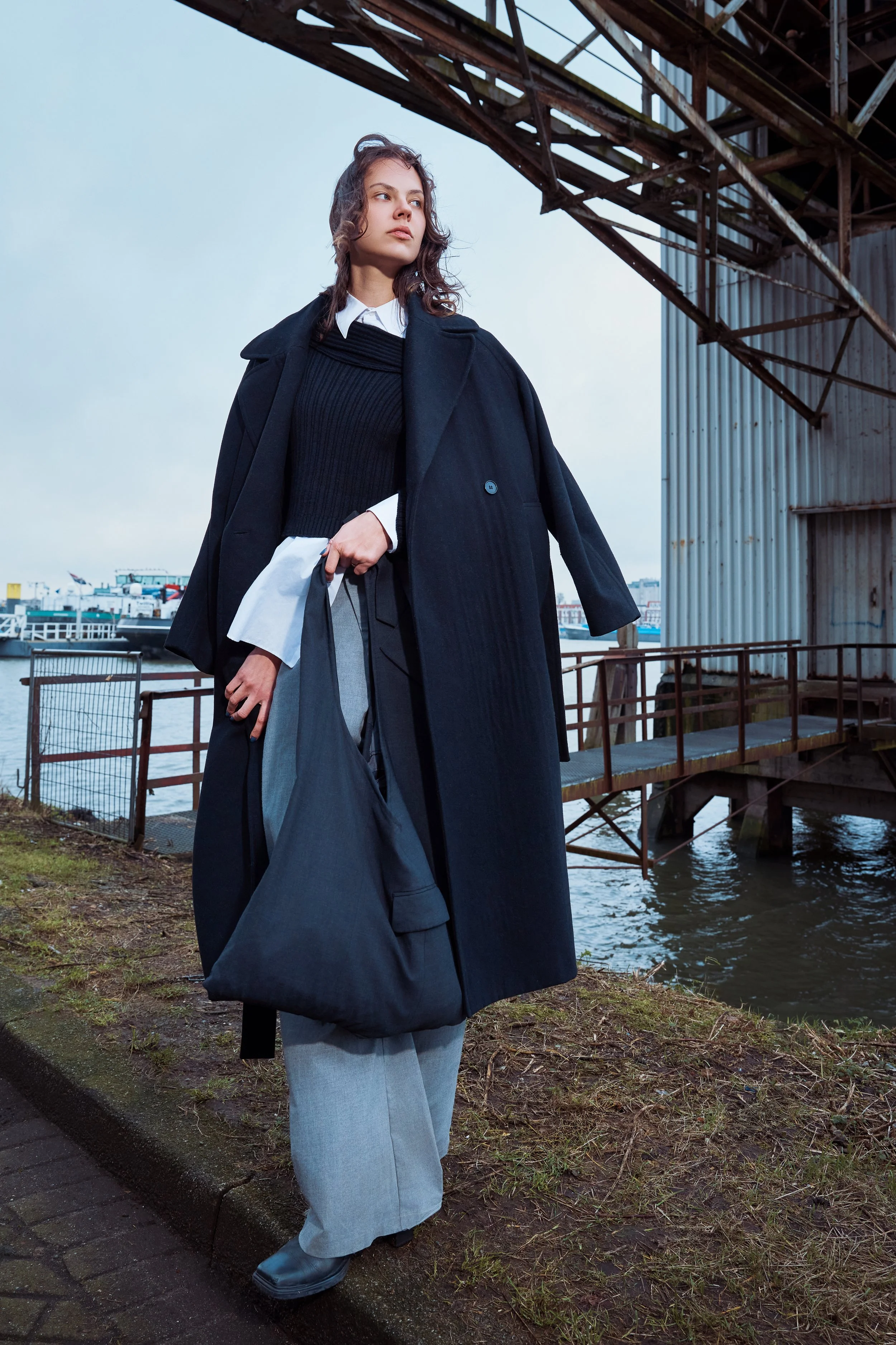A woman standing outdoors near a body of water under a metal structure, wearing a black coat, white shirt, black sweater, gray wide-leg pants, and black shoes, carrying a large black bag.