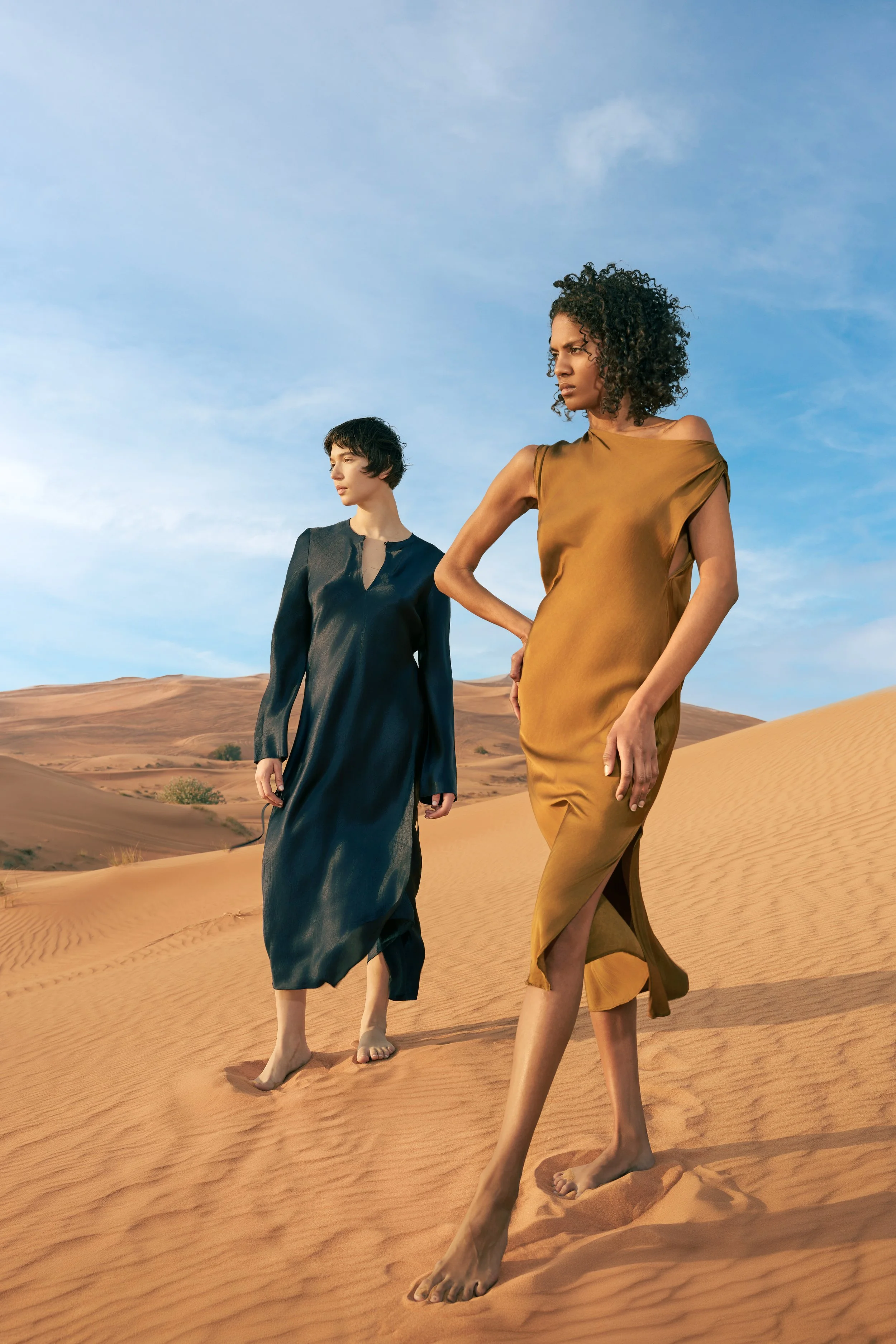 Two women walking barefoot in a desert with sand dunes under a blue sky, wearing elegant dresses.