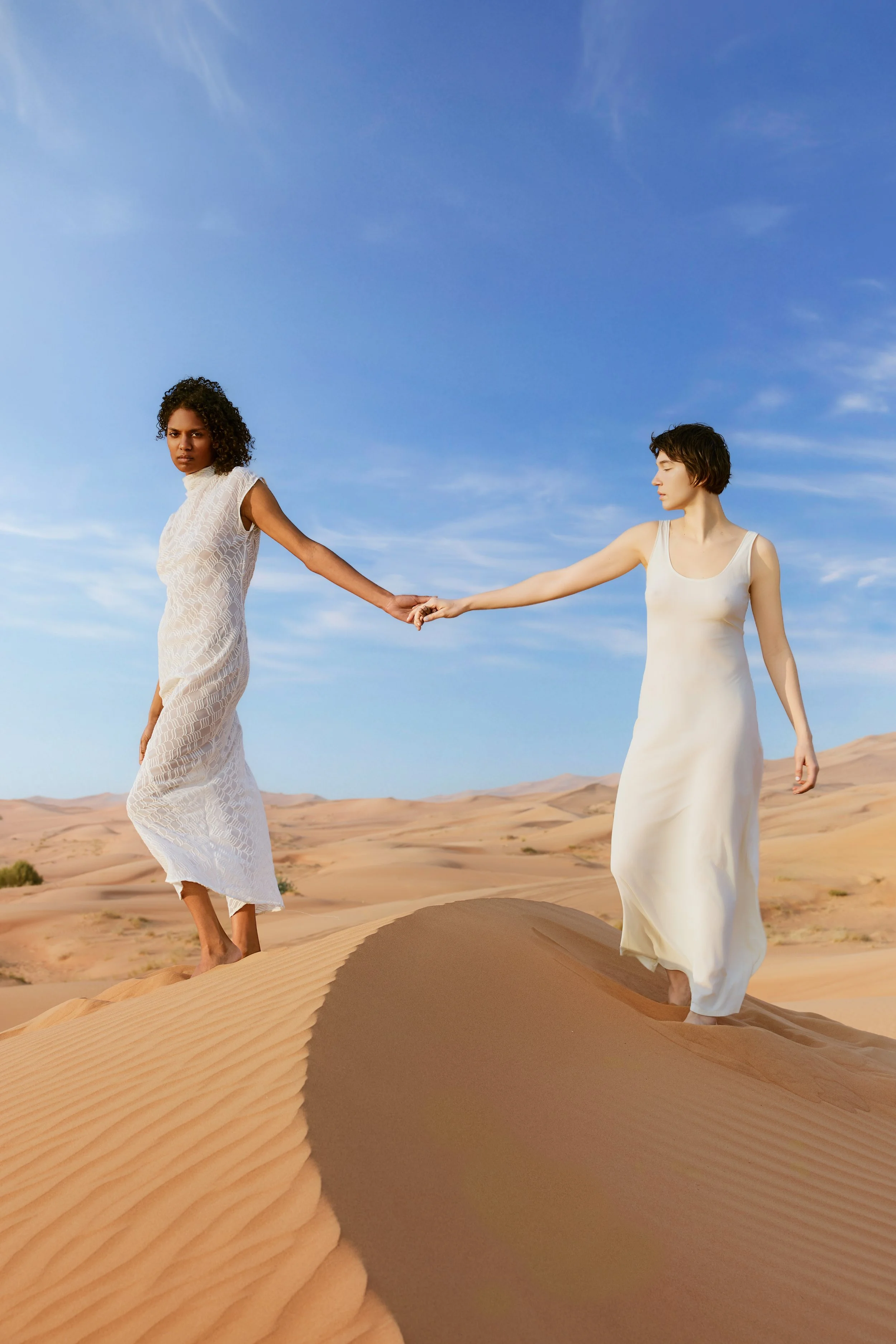 Two women in white dresses walking on sand dunes in a desert, holding hands with a clear blue sky overhead.
