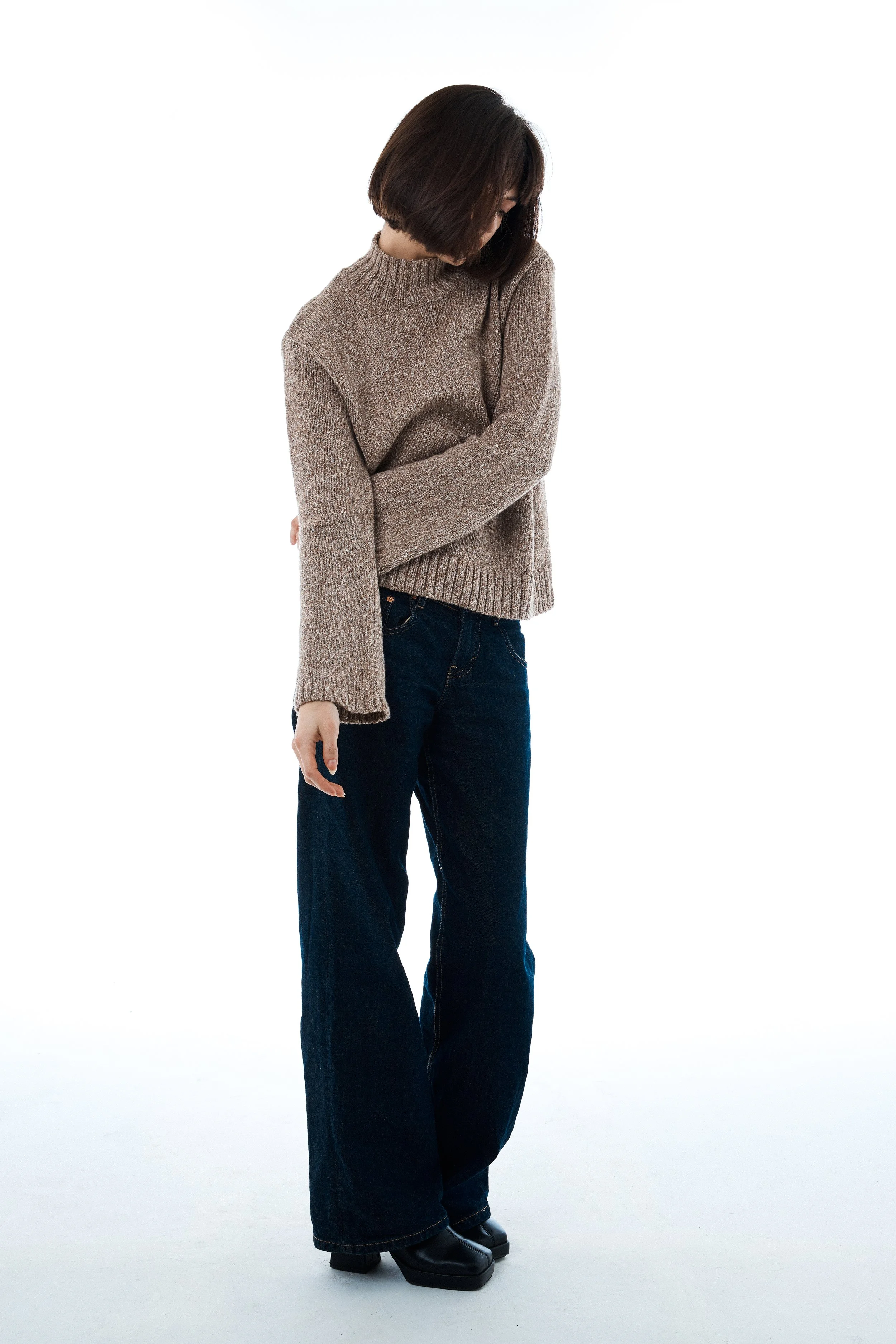 A woman with short brown hair wearing a beige sweater, dark blue jeans, and black boots standing against a plain white background.