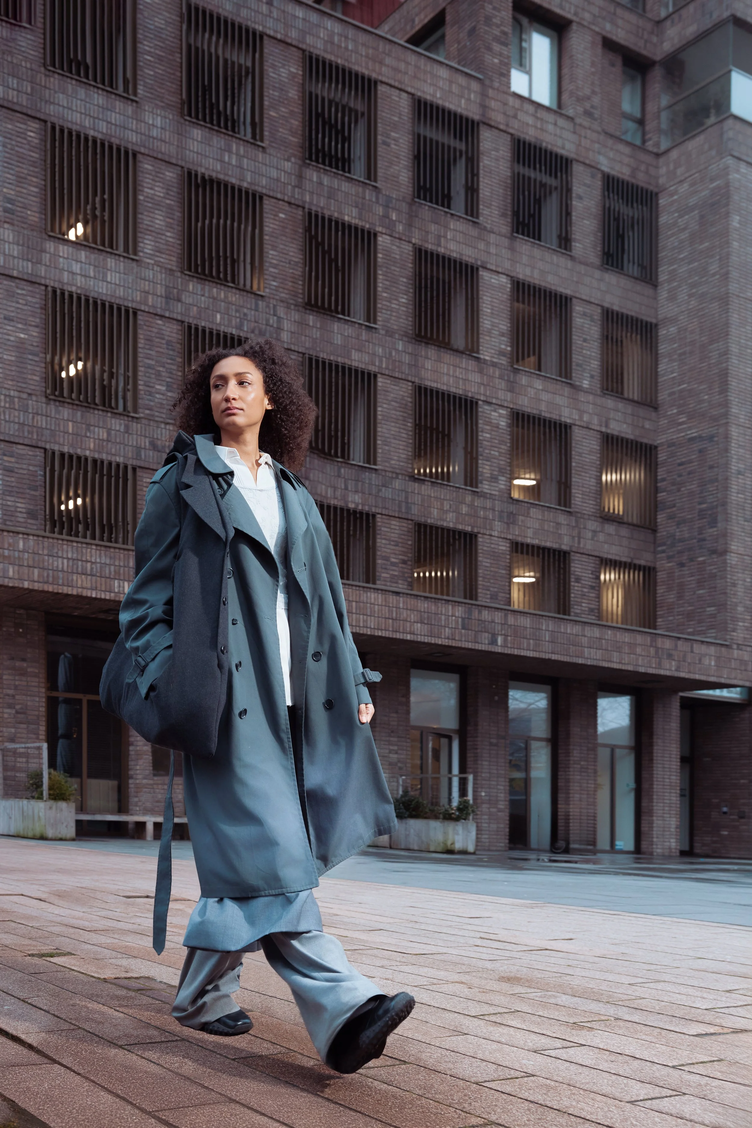 A woman with curly hair walks outside on a rainy day in front of a modern brick building, wearing a long gray trench coat and loose gray pants.