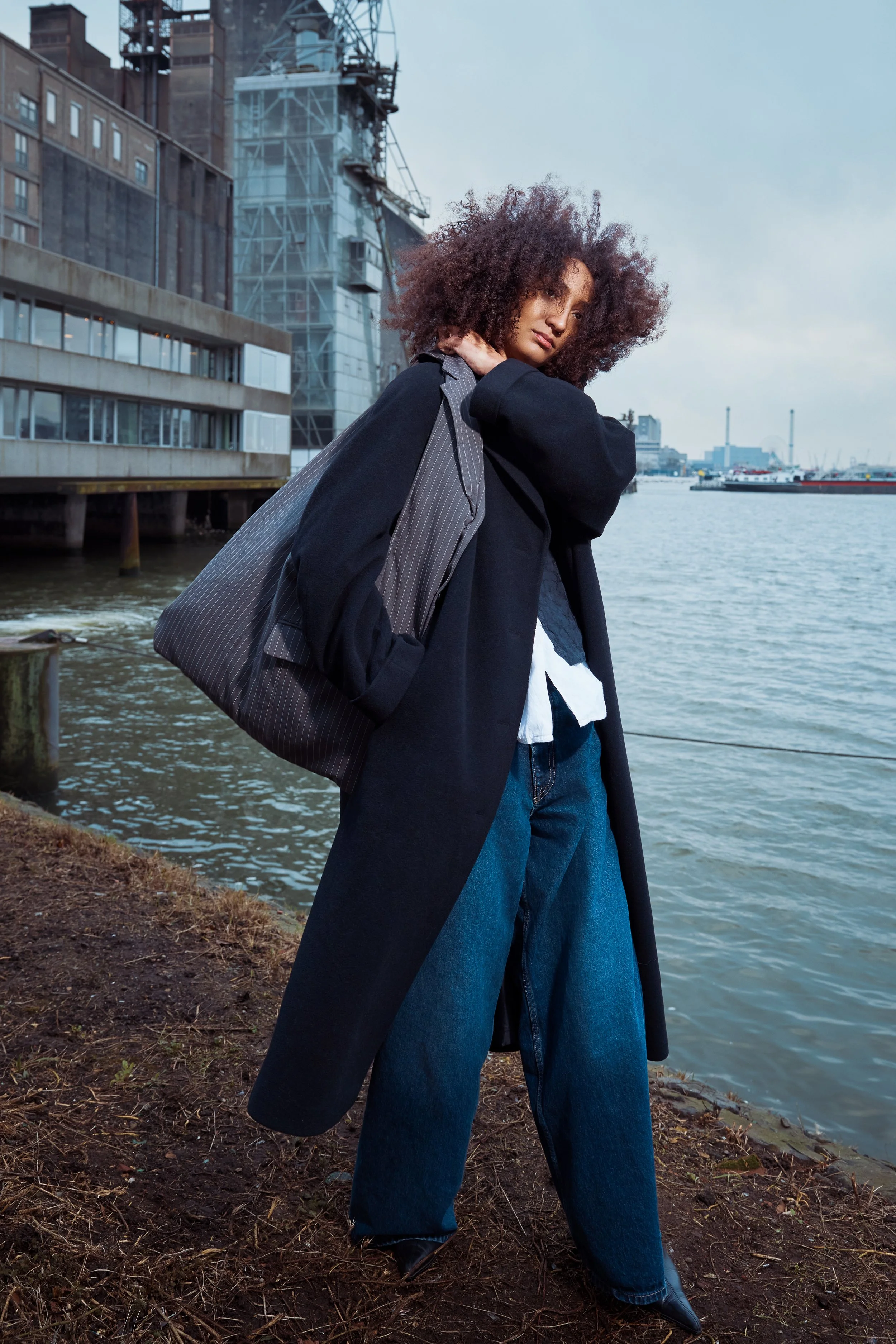 A woman standing near a waterfront with modern buildings and a crane in the background, wearing a black coat, blue jeans, and carrying a striped gray bag over her shoulder.