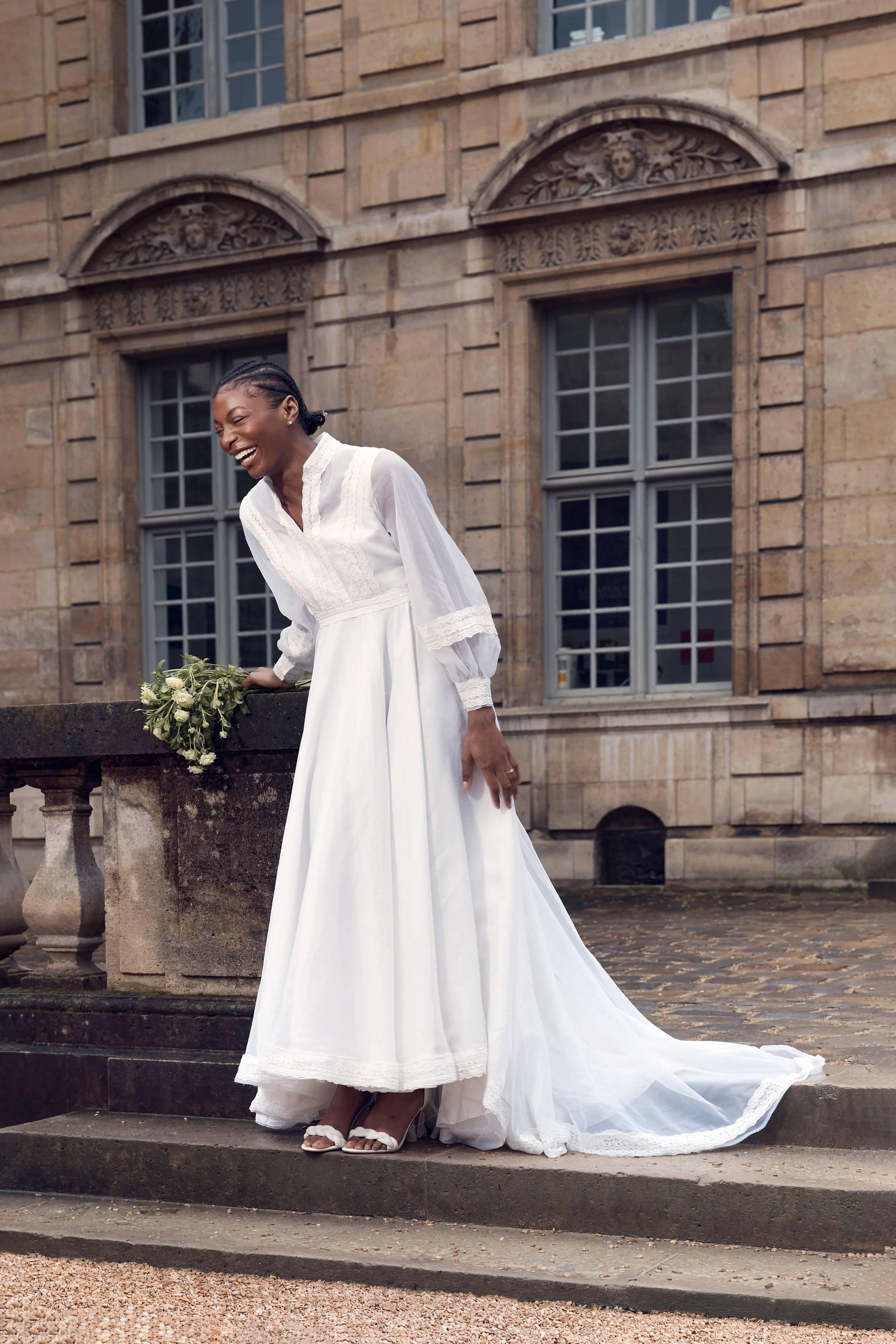 Woman in a white wedding dress smiling and laughing outdoors near a stone building with large windows, holding a bouquet of flowers.