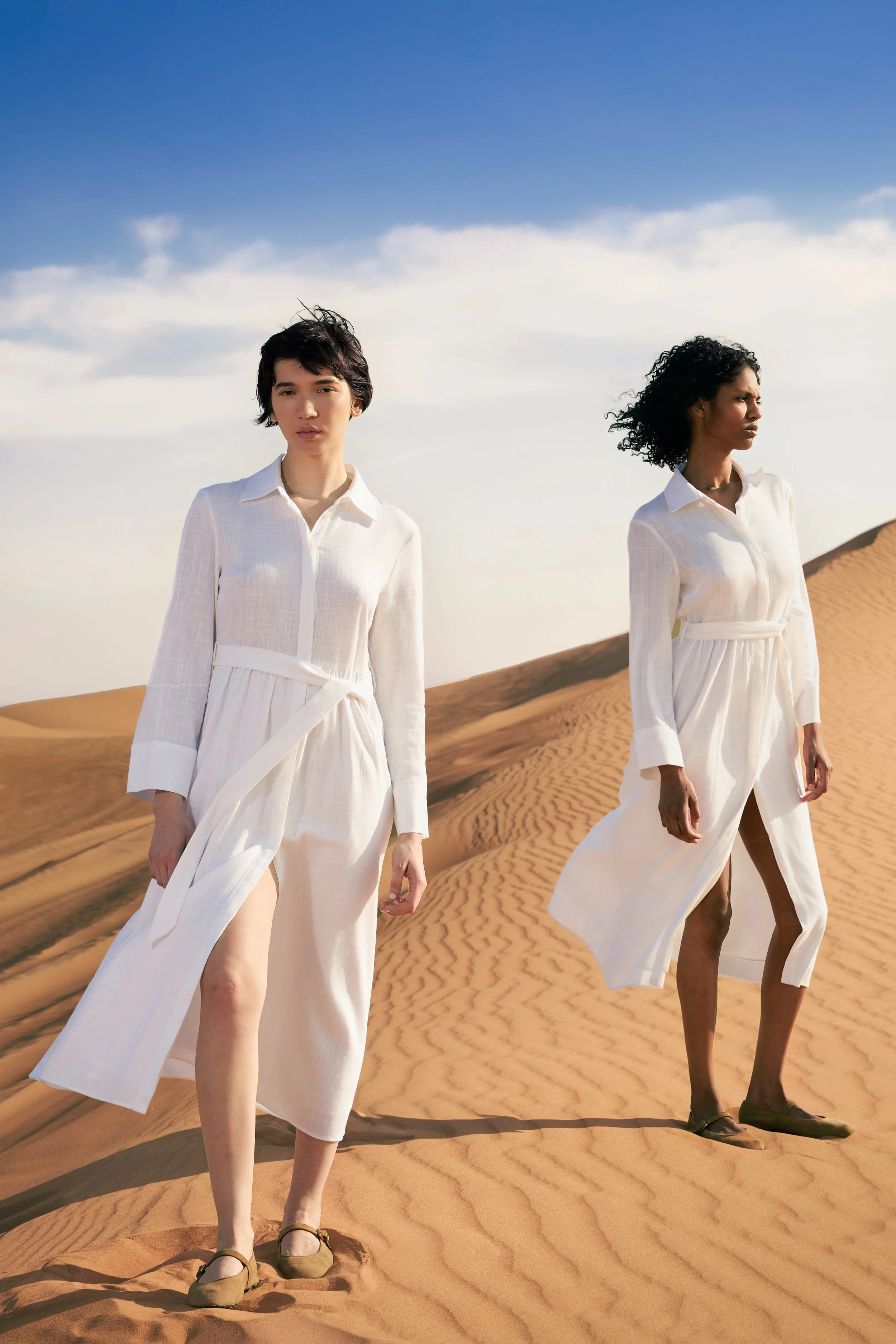Two women in white dresses walking on sand dunes in a desert with a blue sky and clouds in the background.