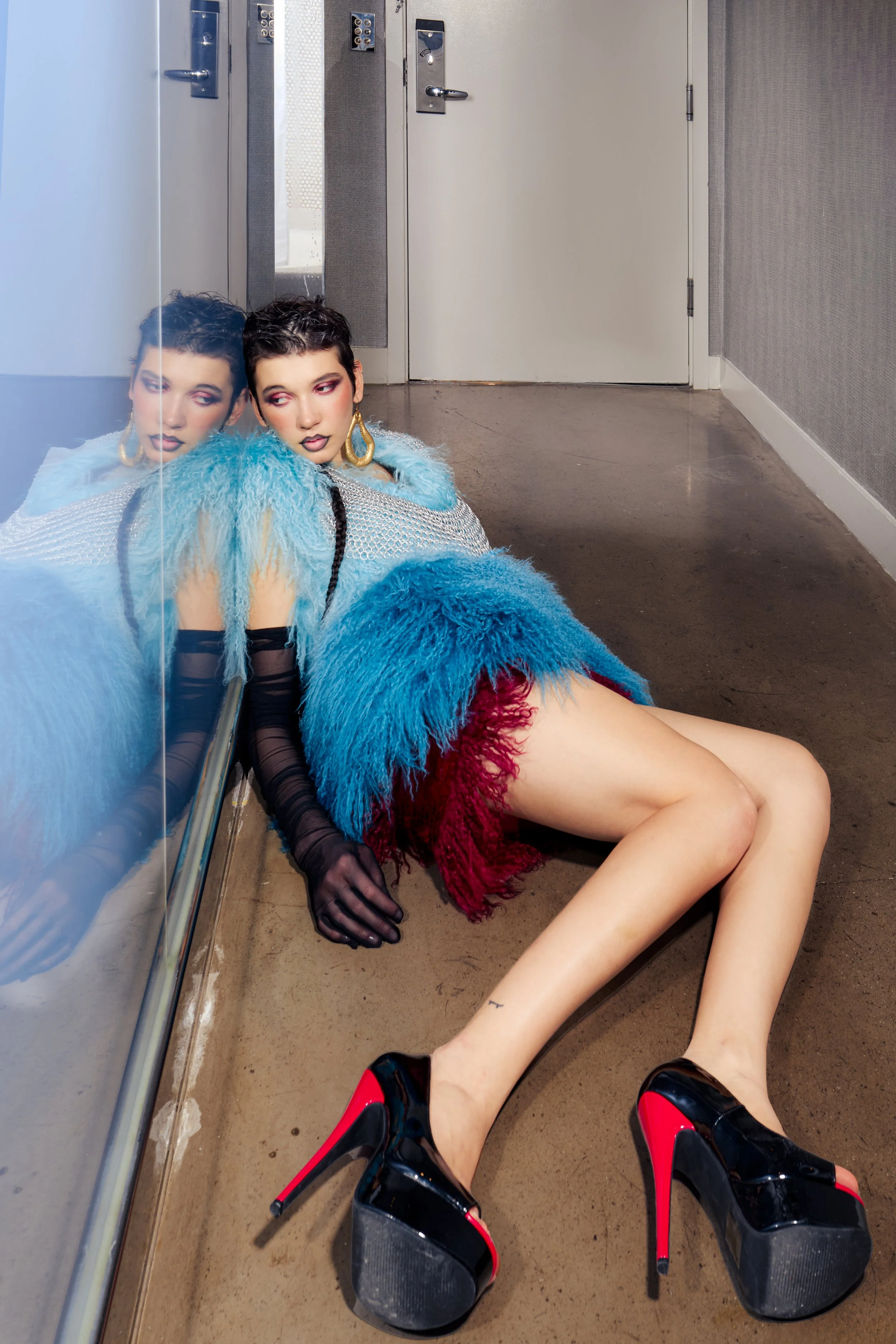 A woman with dark, short hair and bold makeup, including pink eyeshadow and dark lipstick, is sitting on the floor next to a mirror in a hallway. She is wearing a vibrant, multi-colored outfit with a blue and red fuzzy coat, sheer black gloves, and b