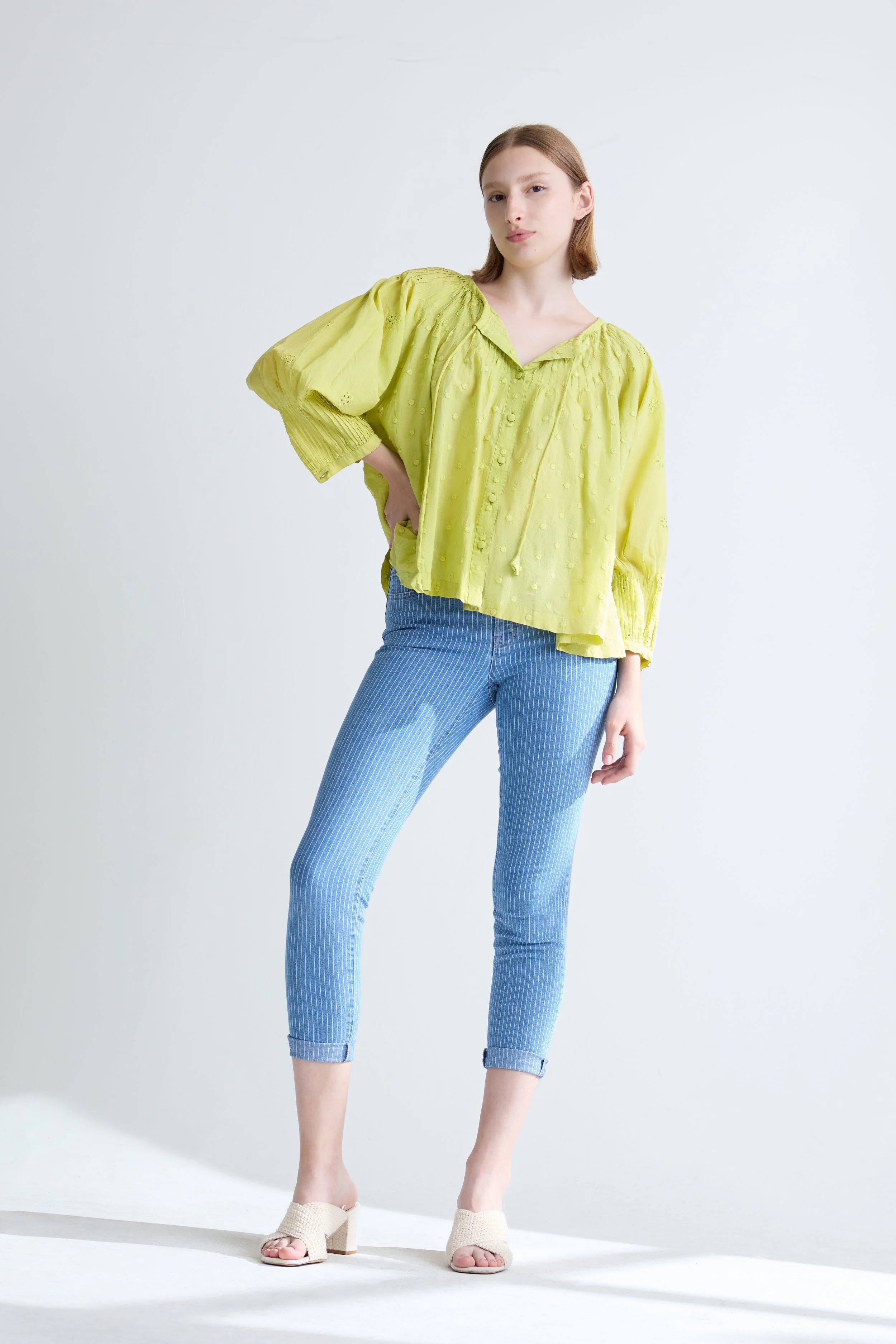 A young woman with reddish-brown hair wearing a bright yellow, loose-fitting, textured blouse with puffed sleeves, paired with light blue, pinstriped jeans and beige heeled sandals, standing against a plain white background.