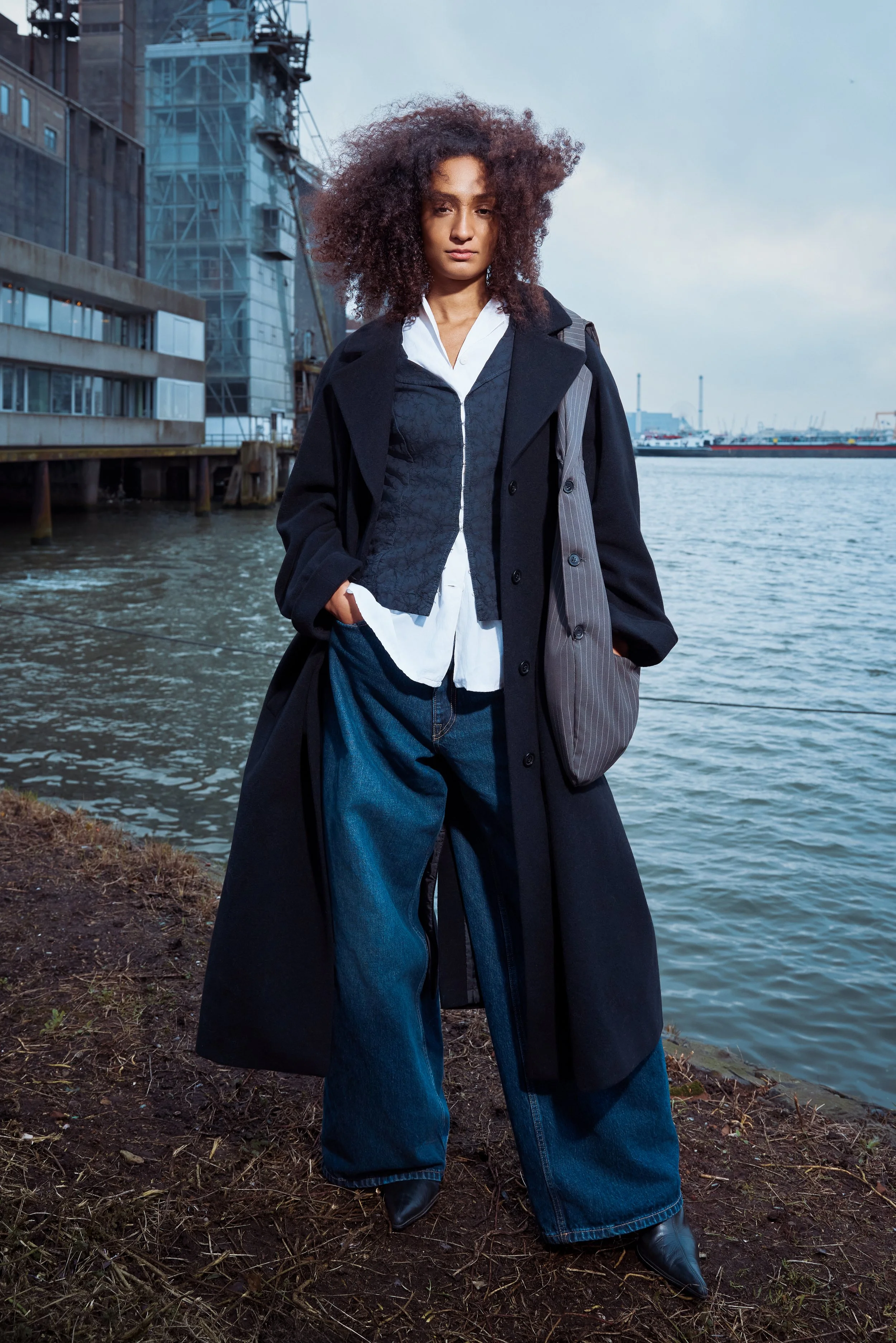 A woman standing by a waterfront, wearing a layered outfit with a long black coat, wide-leg jeans, a white shirt, and a black vest, with curly hair and a serious expression, in an urban area near water.