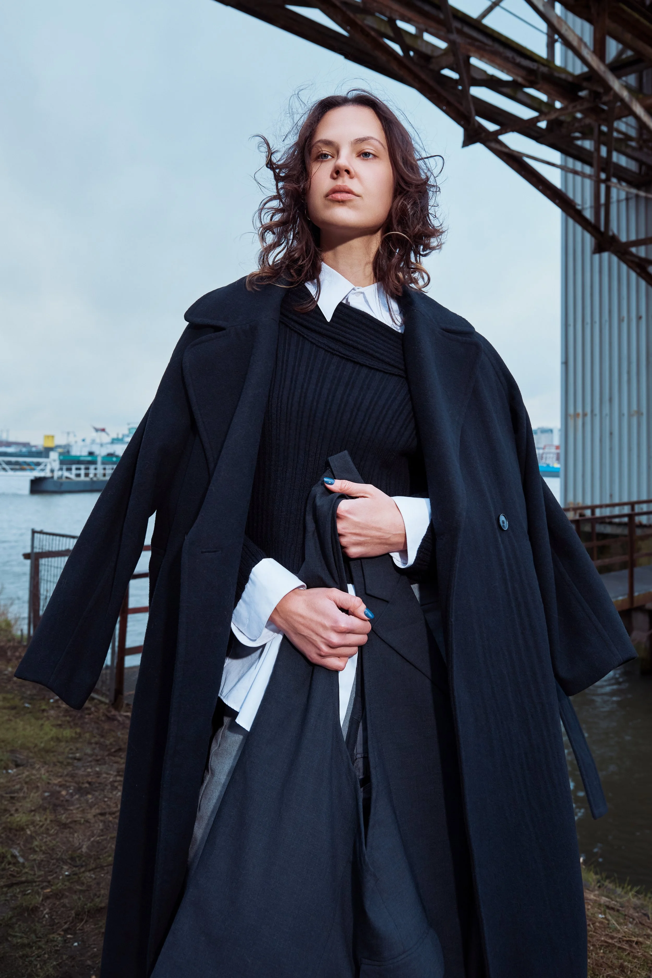 A woman dressed in layered professional black and white clothing, standing outdoors near water with a bridge in the background.