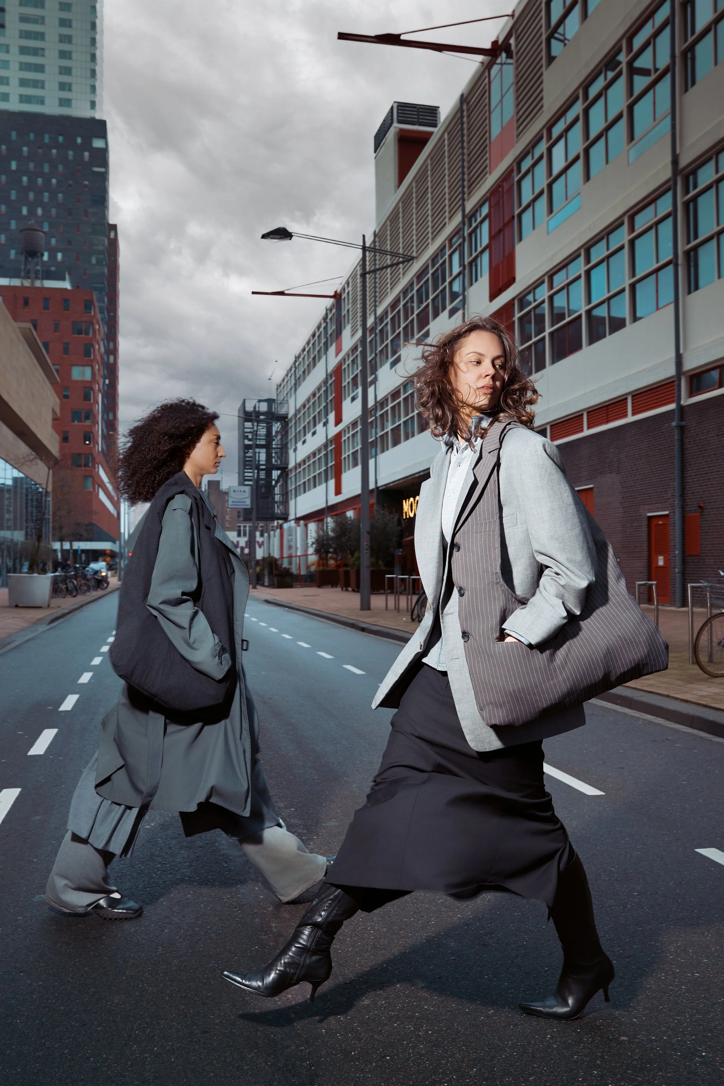 Two women walking across an urban street with modern buildings in the background, under a cloudy sky.