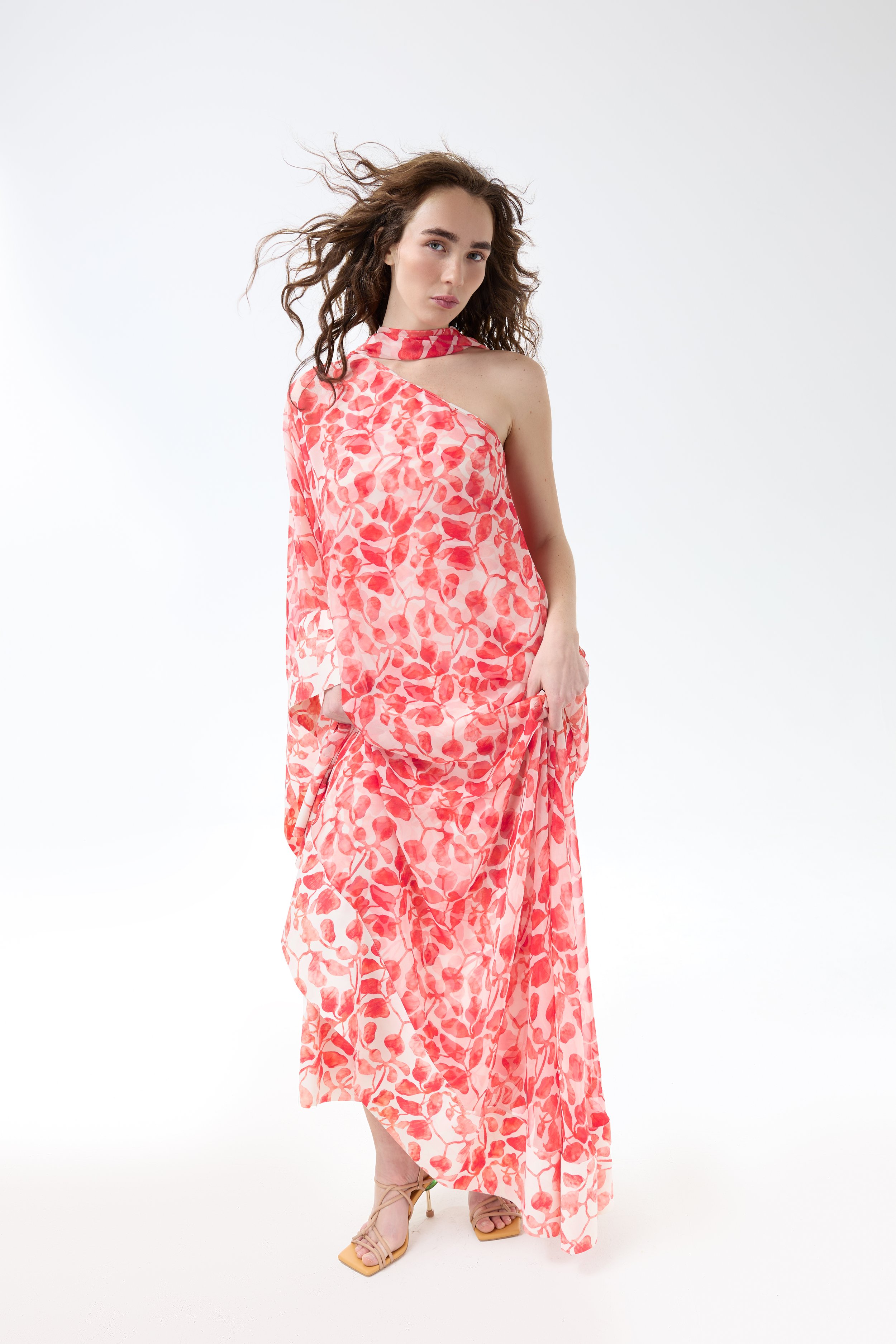A woman with curly brown hair wearing a pink and red floral dress, standing against a plain white background.