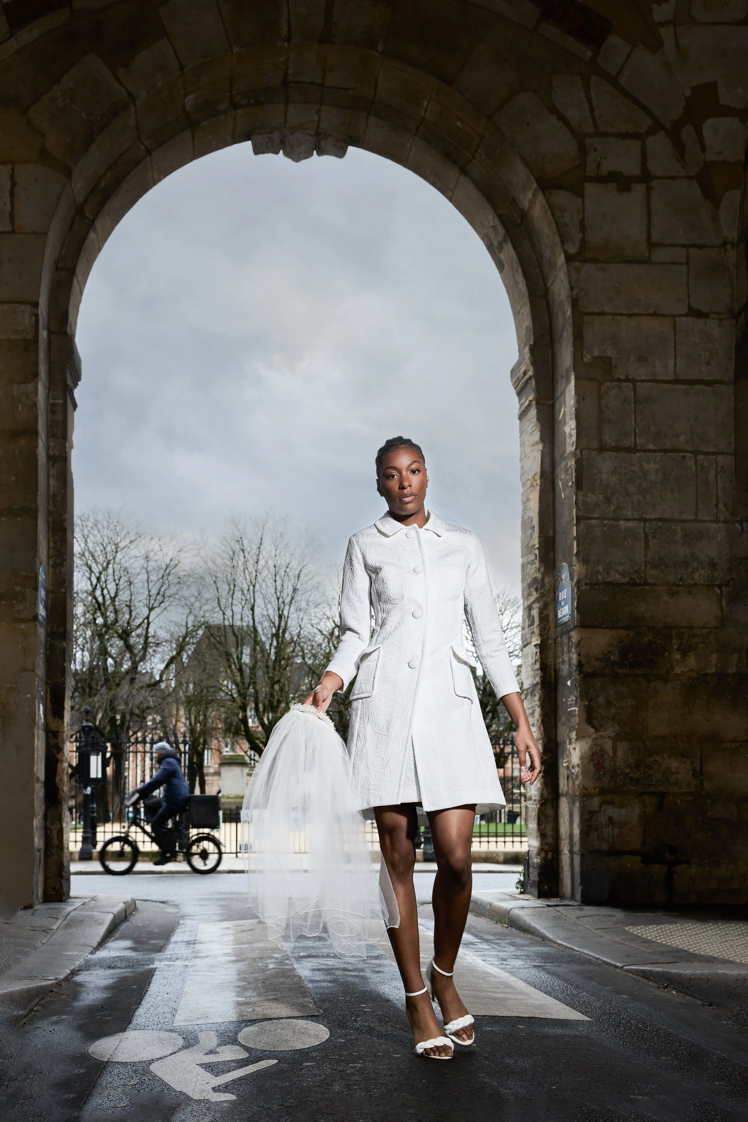 A woman dressed in a white coat and high heels walking through a stone archway with a cloudy sky and trees in the background, holding a white tulle veil or dress.