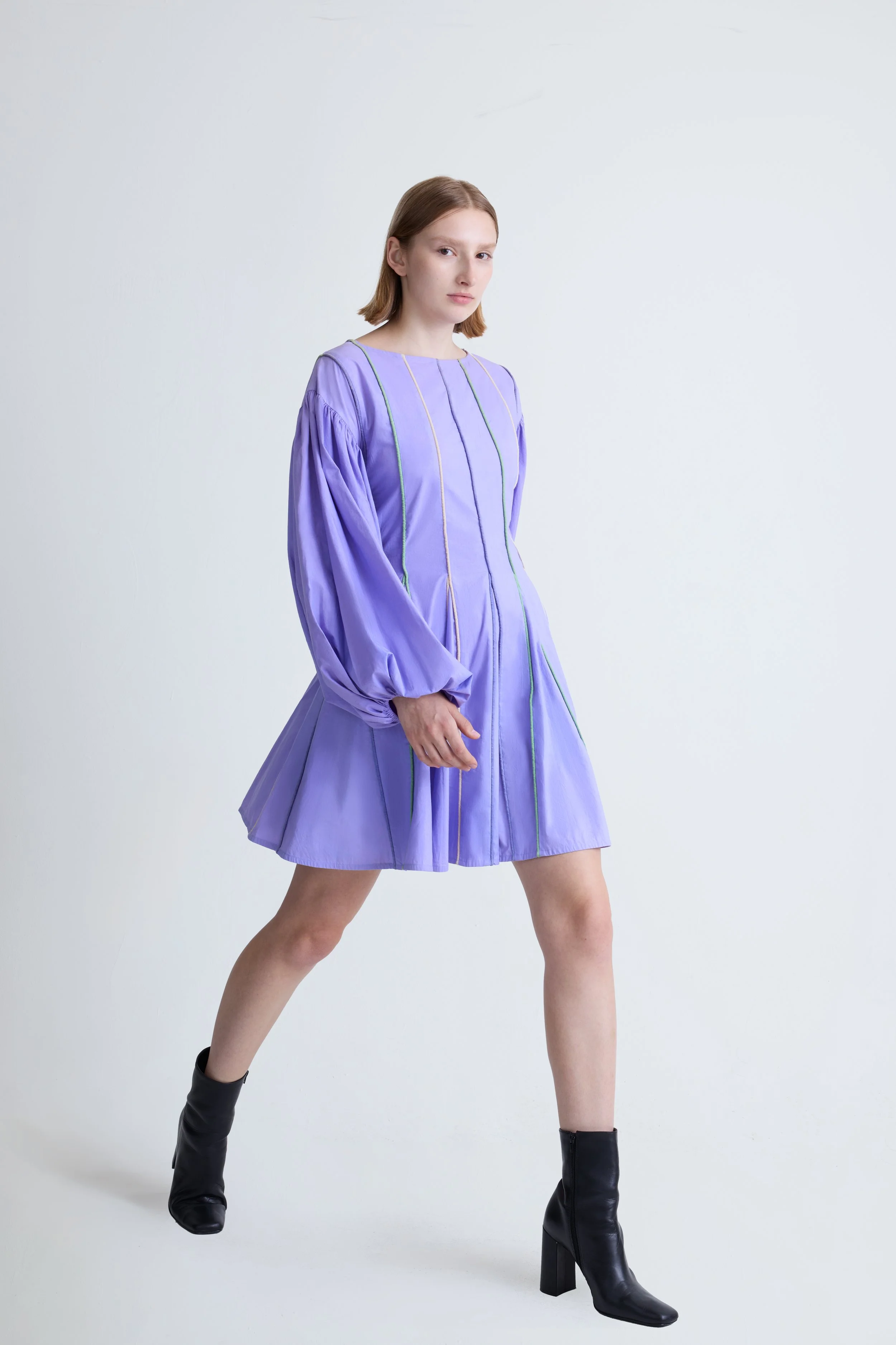 A young woman with straight, shoulder-length red hair wearing a lavender dress with long, puffy sleeves and multicolored vertical stitching, paired with black heeled boots, standing against a plain white background.