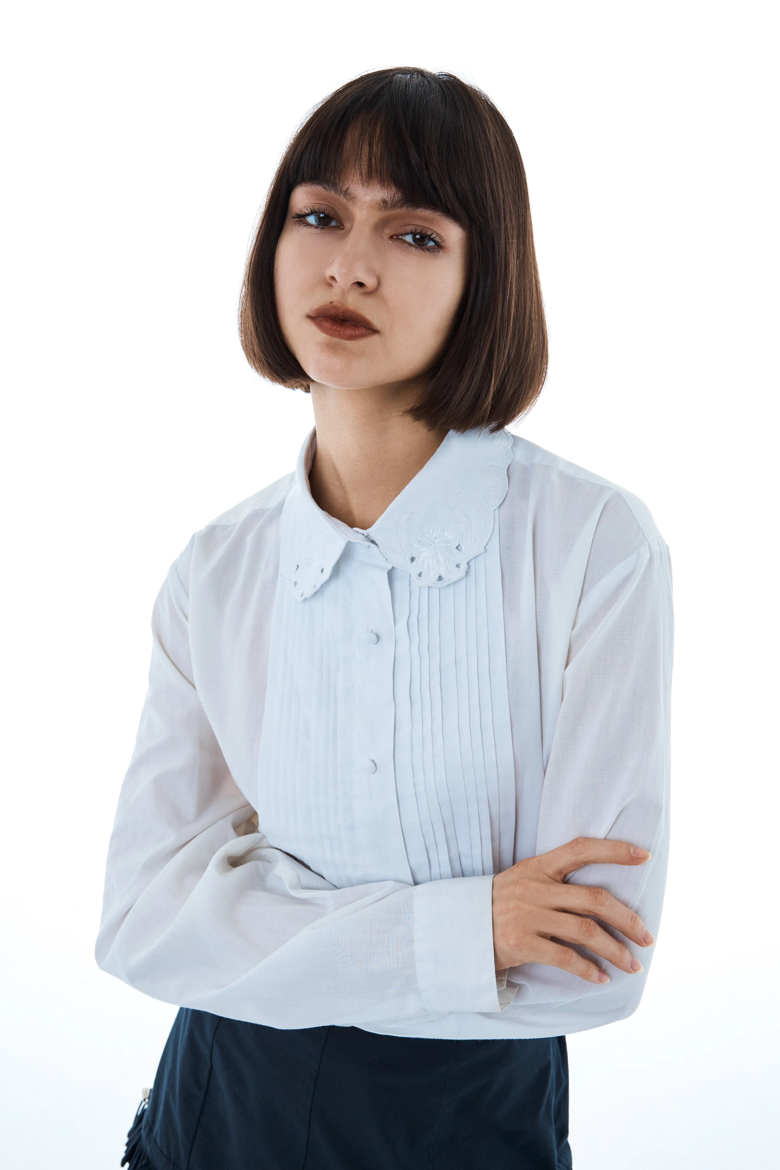 A young woman with shoulder-length dark hair and bangs, wearing a white blouse with a Peter Pan collar and plum-colored lipstick, crosses her arms and looks directly at the camera with a neutral expression.