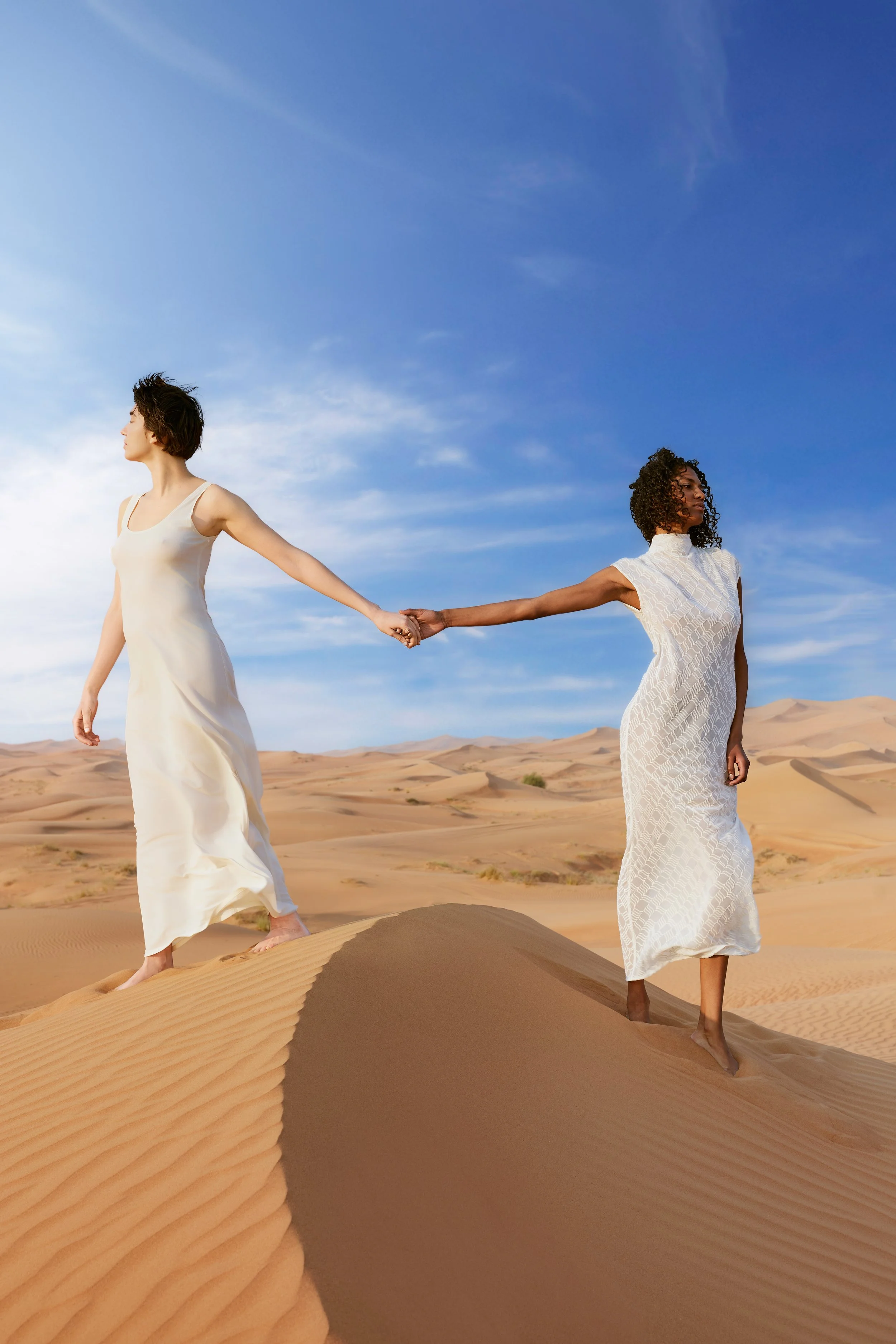 Two women in white dresses holding hands in a desert landscape with sand dunes and a blue sky.