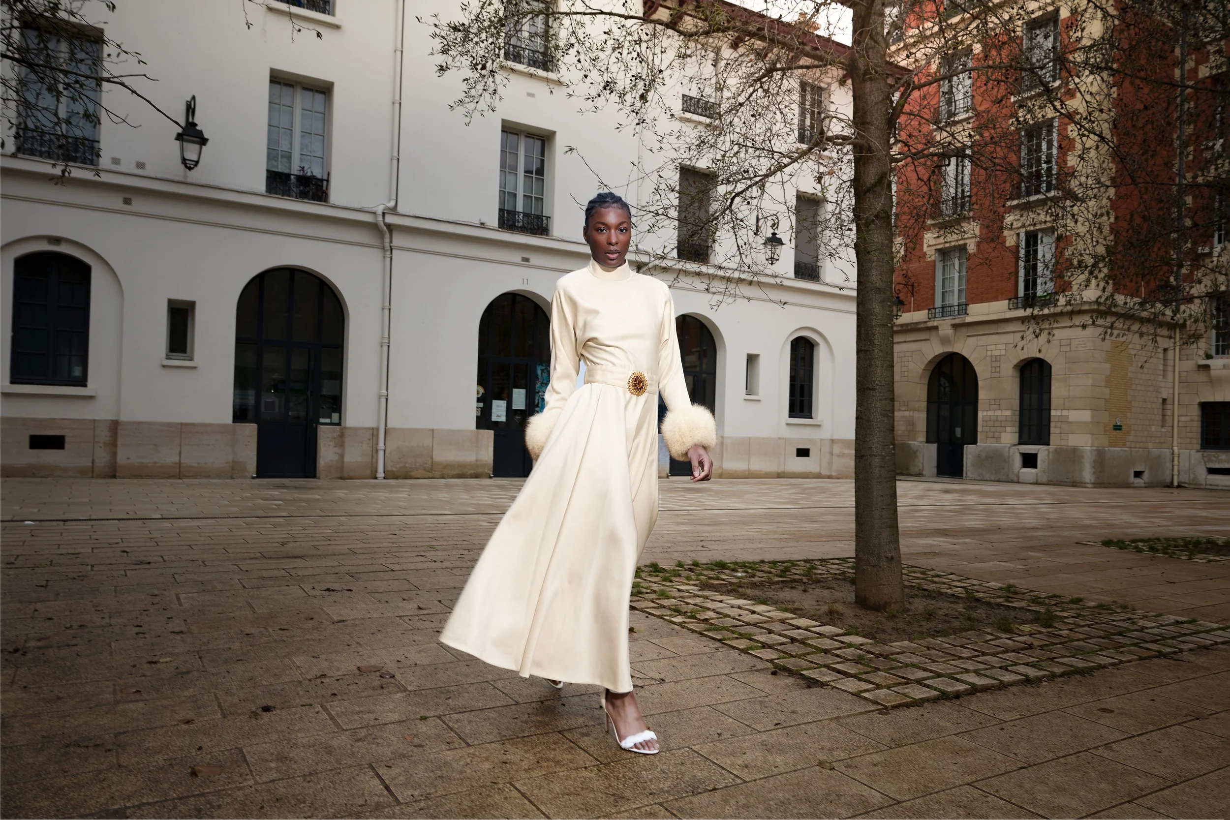 A woman in a long, elegant cream-colored dress with fur cuffs and a decorative belt stands on a paved courtyard with residential buildings in the background. She is wearing white high-heeled sandals and has her hair styled up.