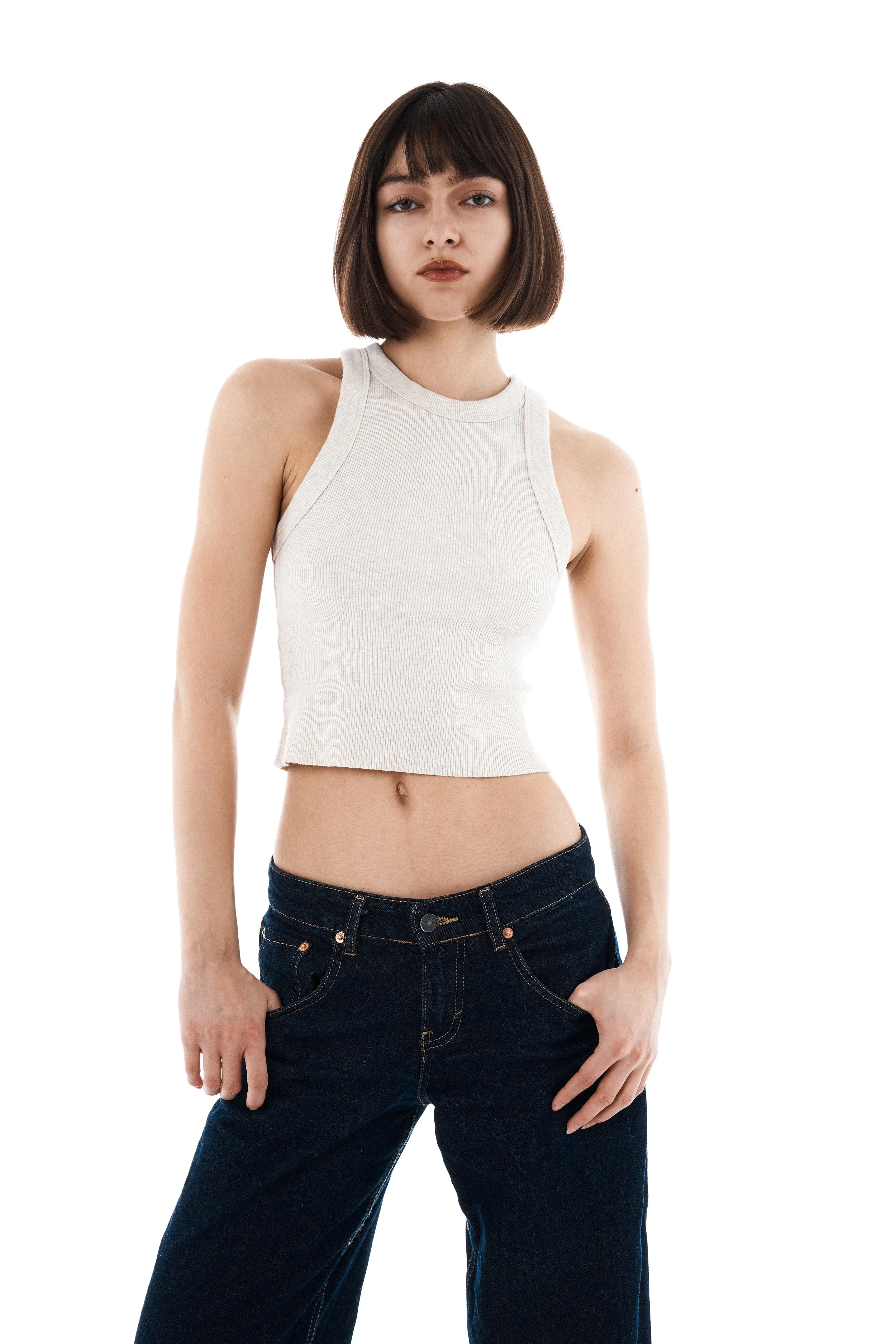 Young woman with dark brown hair and bangs posing against a white background, wearing a sleeveless off-white crop top and dark jeans.