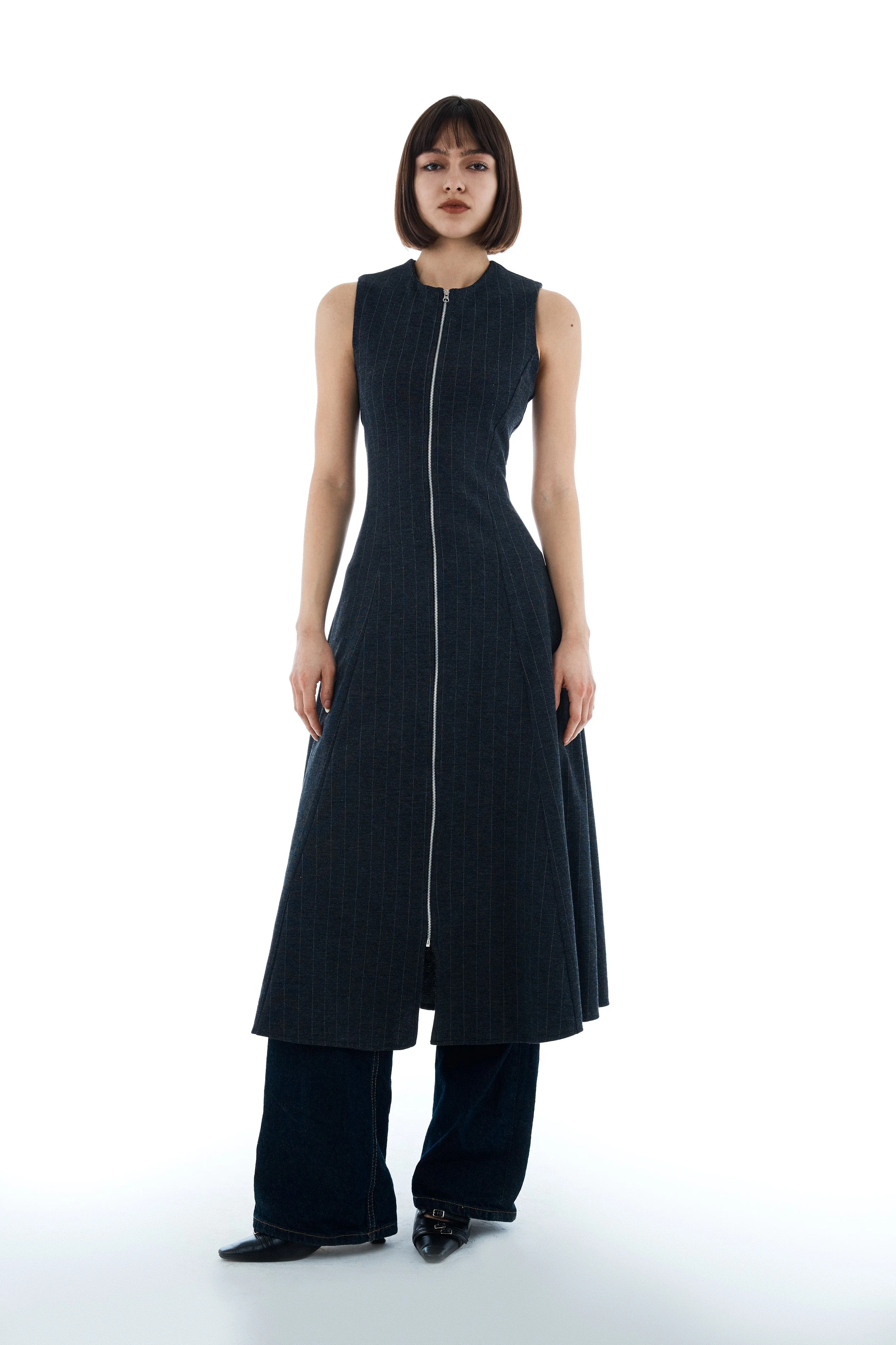 Young woman with bob haircut wearing a sleeveless dark blue dress with vertical stripes and a front zipper, paired with wide-leg jeans and pointed shoes, standing against a plain white background.