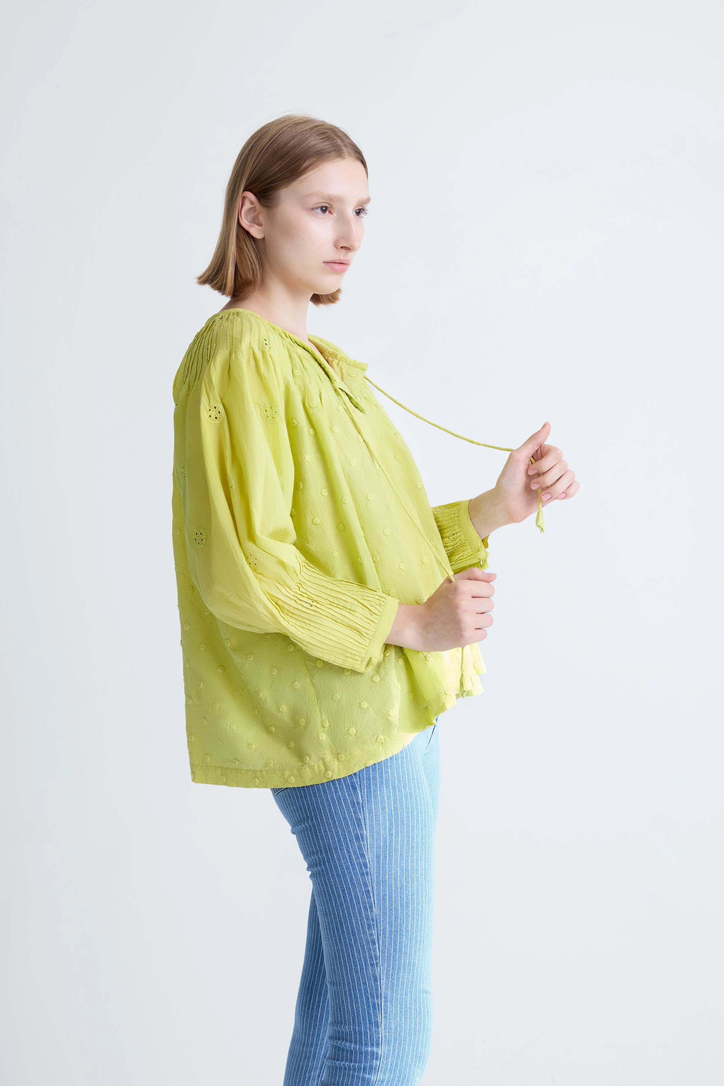 A young woman with shoulder-length light brown hair wearing a loose, bright yellow blouse and light blue striped pants, standing against a plain white background.