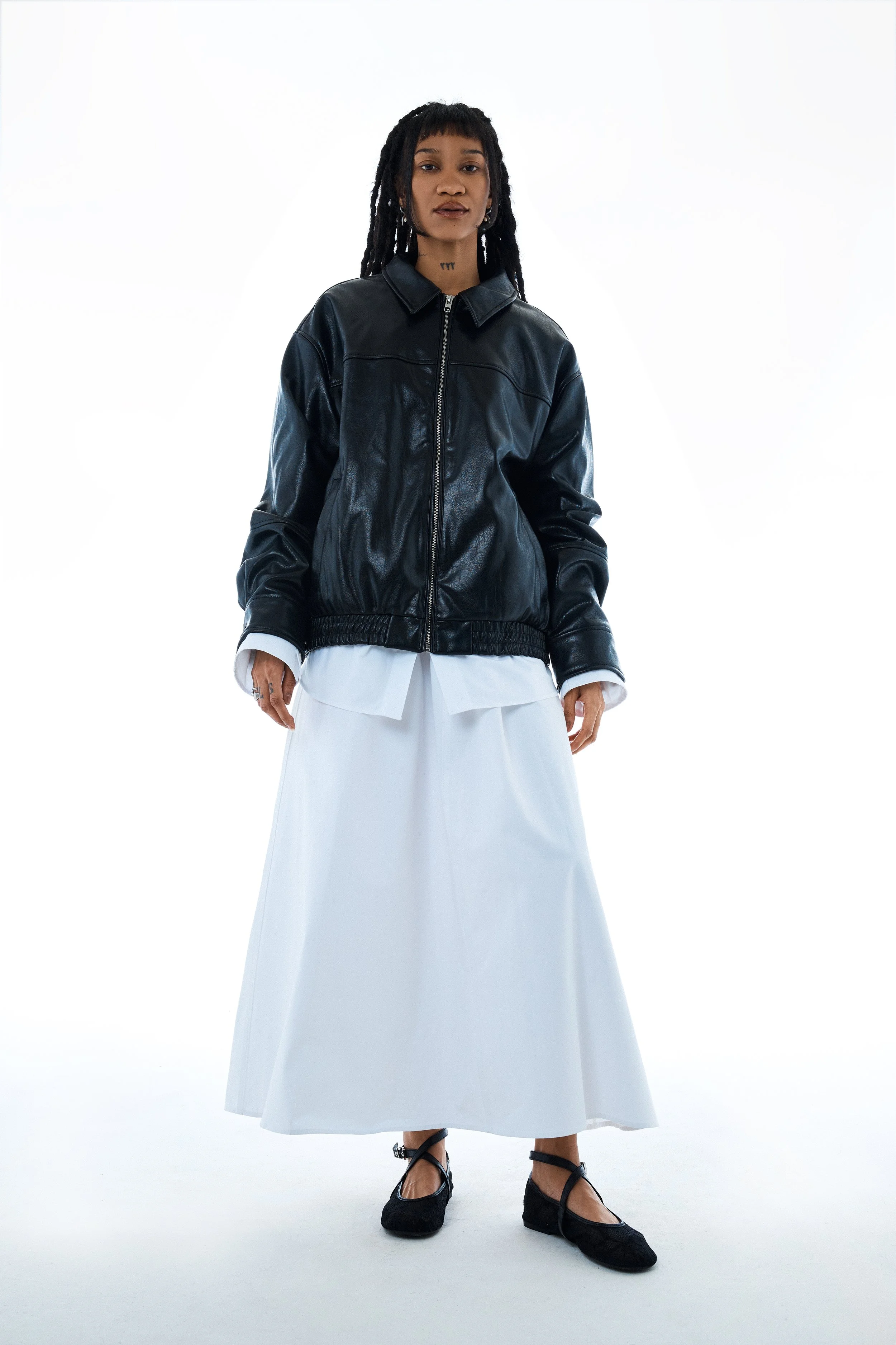 A woman posing in a studio with a white background, wearing a black leather jacket, a white long shirt, a long white skirt, and black shoes.