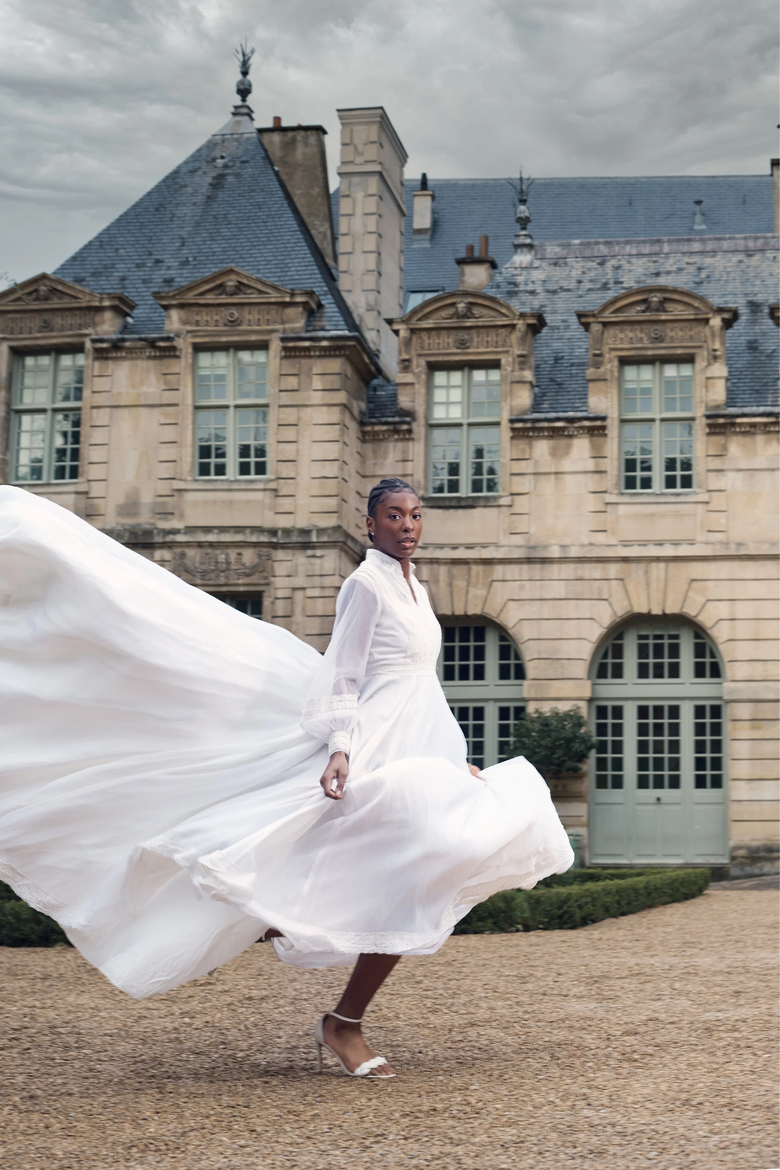 A woman wearing a long white dress and heels walking in front of a historic stone building with multiple windows and ornate roof details.