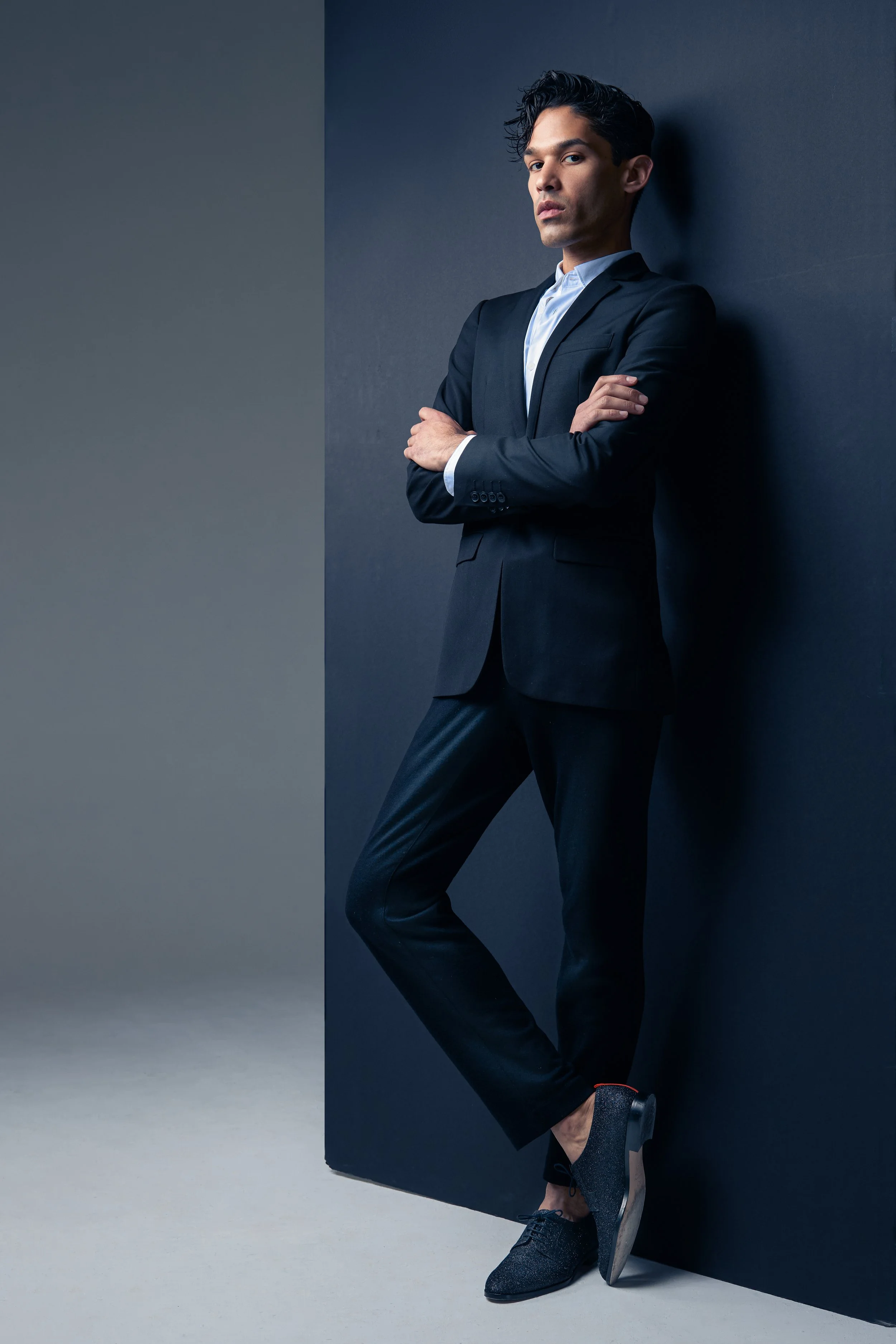 A man in a black suit with a white shirt, standing with arms crossed, leaning against a dark blue wall in a studio setting.