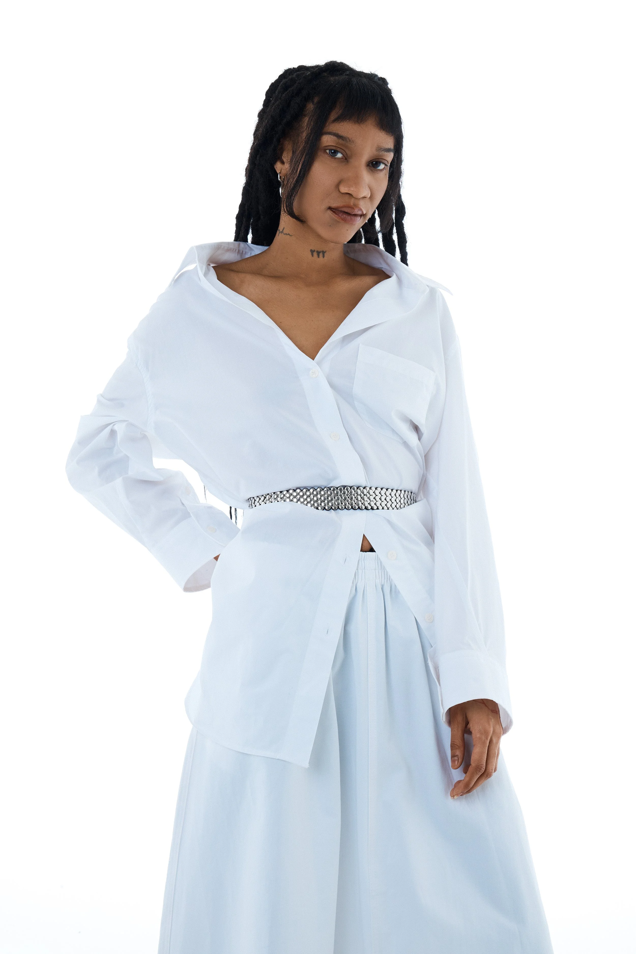 Woman with dreadlocks wearing a white, button-up shirt with rolled-up sleeves, a silver belt, and wide-leg white pants, standing against a white background.