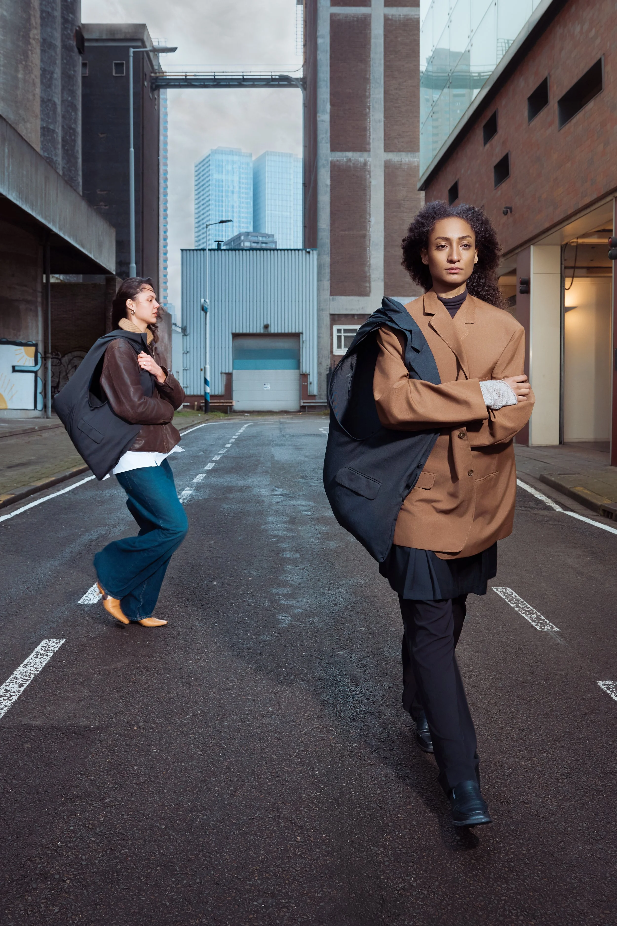 Two women walking on an urban street with tall buildings in the background. The woman in front is crossing her arms, wearing a brown blazer, black pants, and carrying a backpack. The woman behind is standing with one foot slightly raised, wearing a l